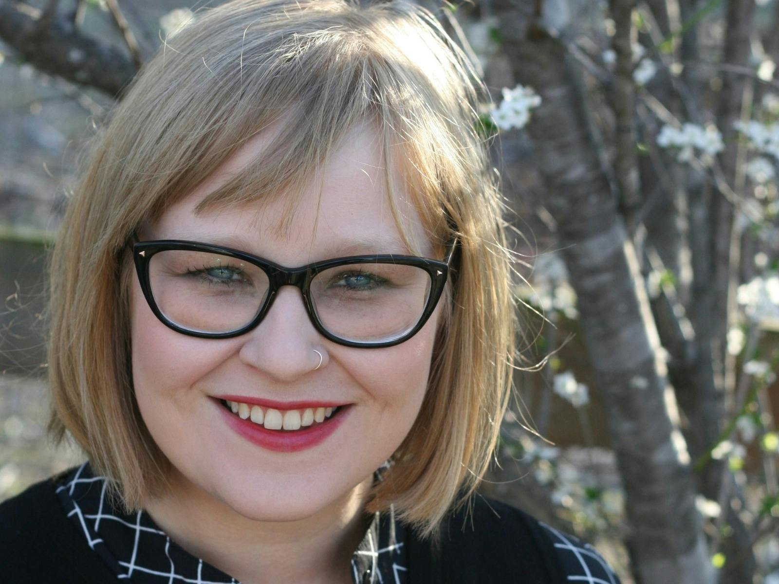 A smiling woman with glasses and blonde hair stands outdoors near blooming trees.