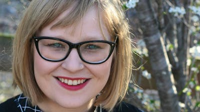 A smiling woman with glasses and blonde hair stands outdoors near blooming trees.