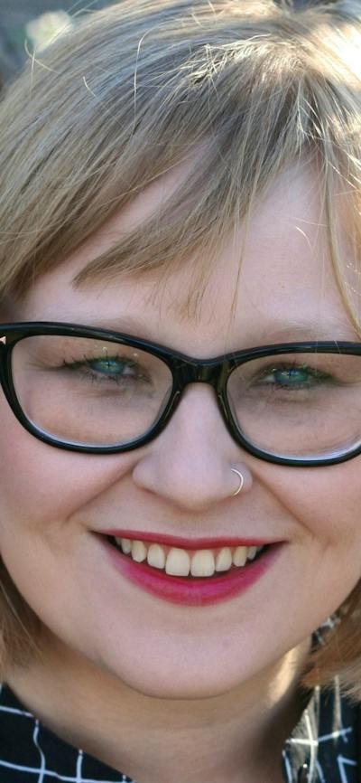 A smiling woman with glasses and blonde hair stands outdoors near blooming trees.