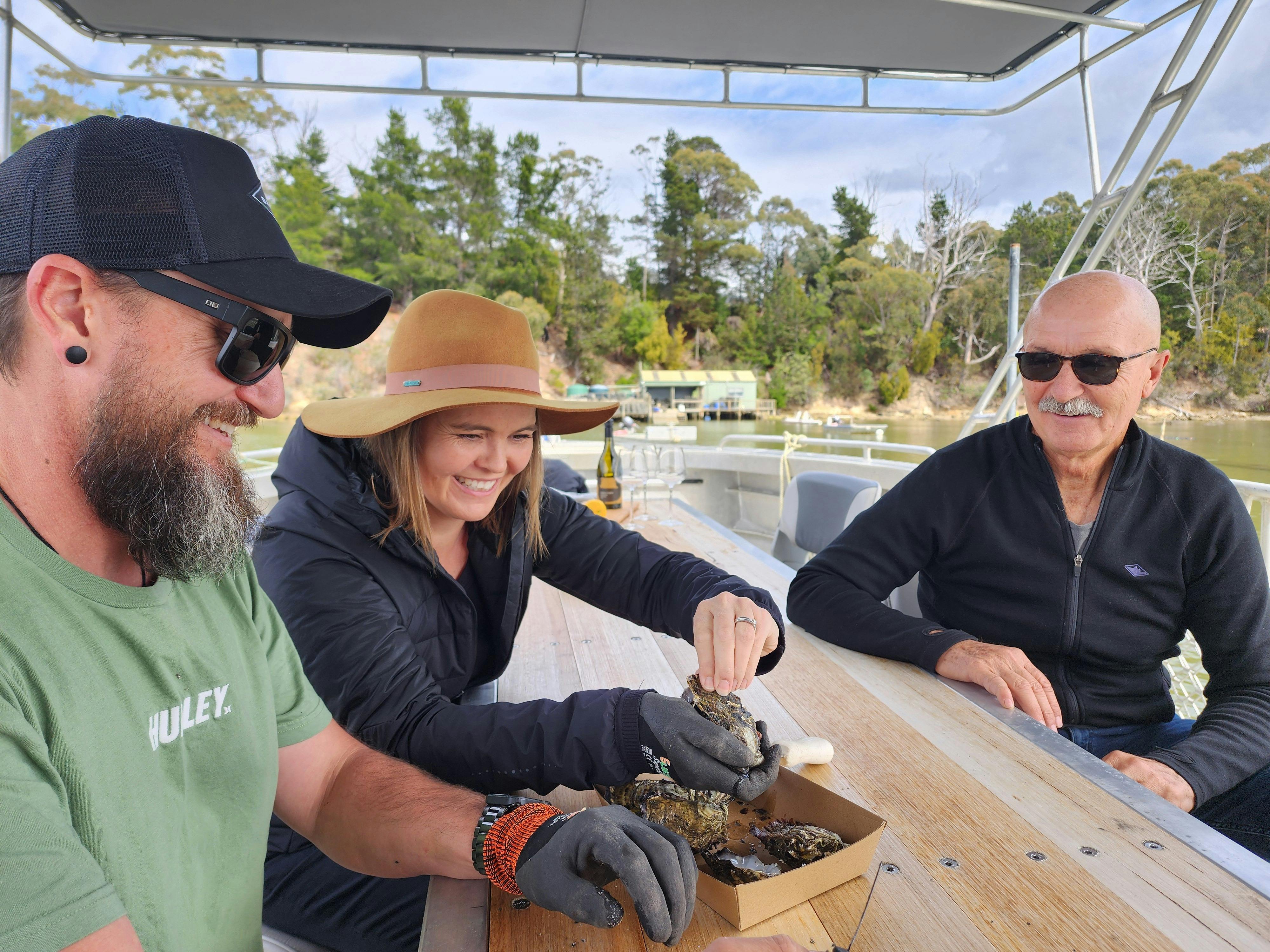 People shucking oysters and enjoying wine aboard boat; water and forested shore in background