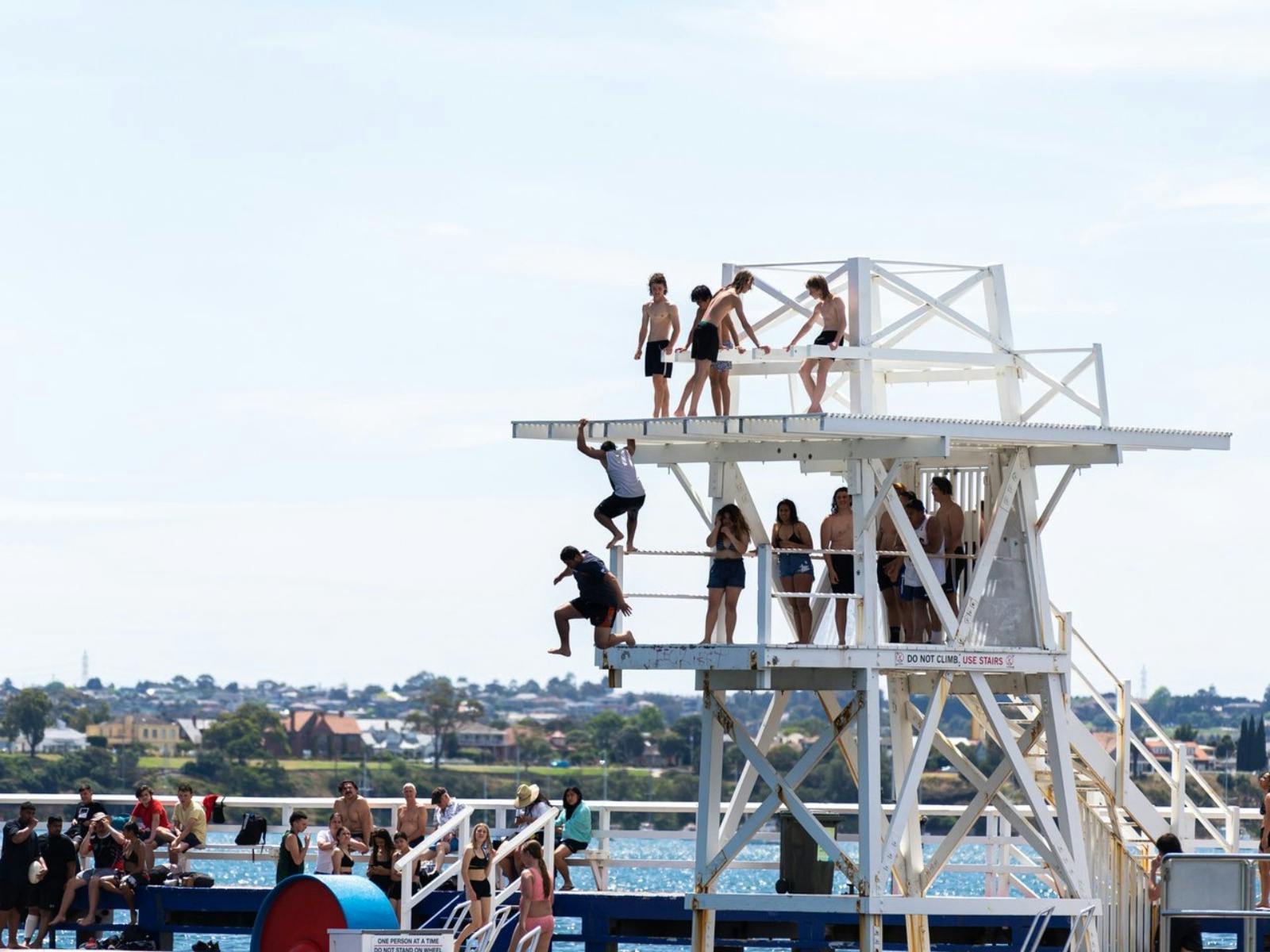 Many youthful people lining up to jump off diving board into Eastern Beach swimming reserve