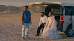 A bride and groom sit in the back of an open van in the desert. Another man stands beside them.