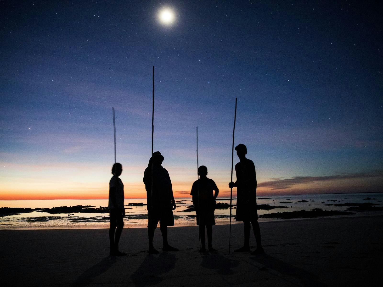 4 silhouettes standing with spears and the sunset behind them.