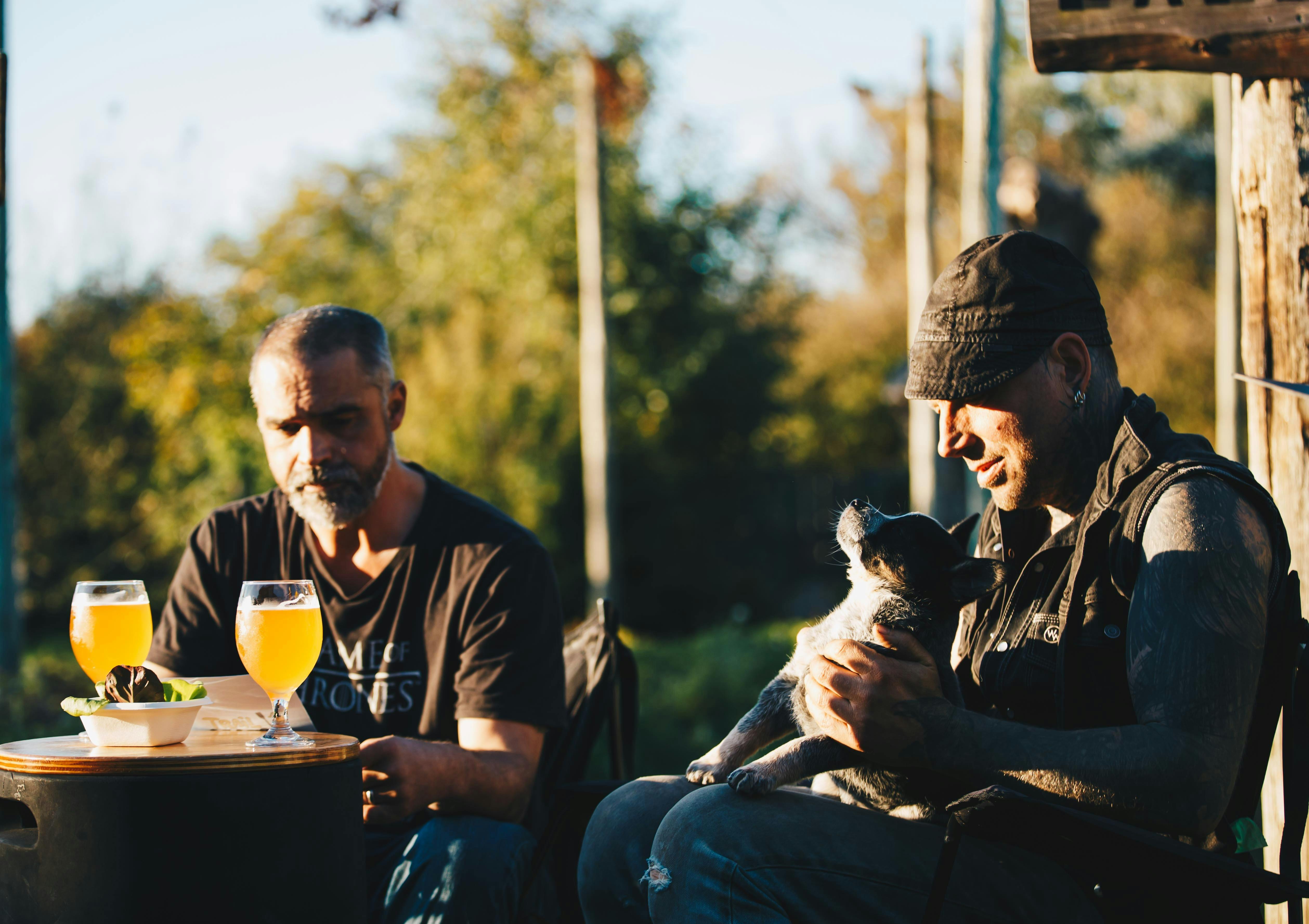 Two men sitting outside with glasses of beer and a blue heeler puppy.