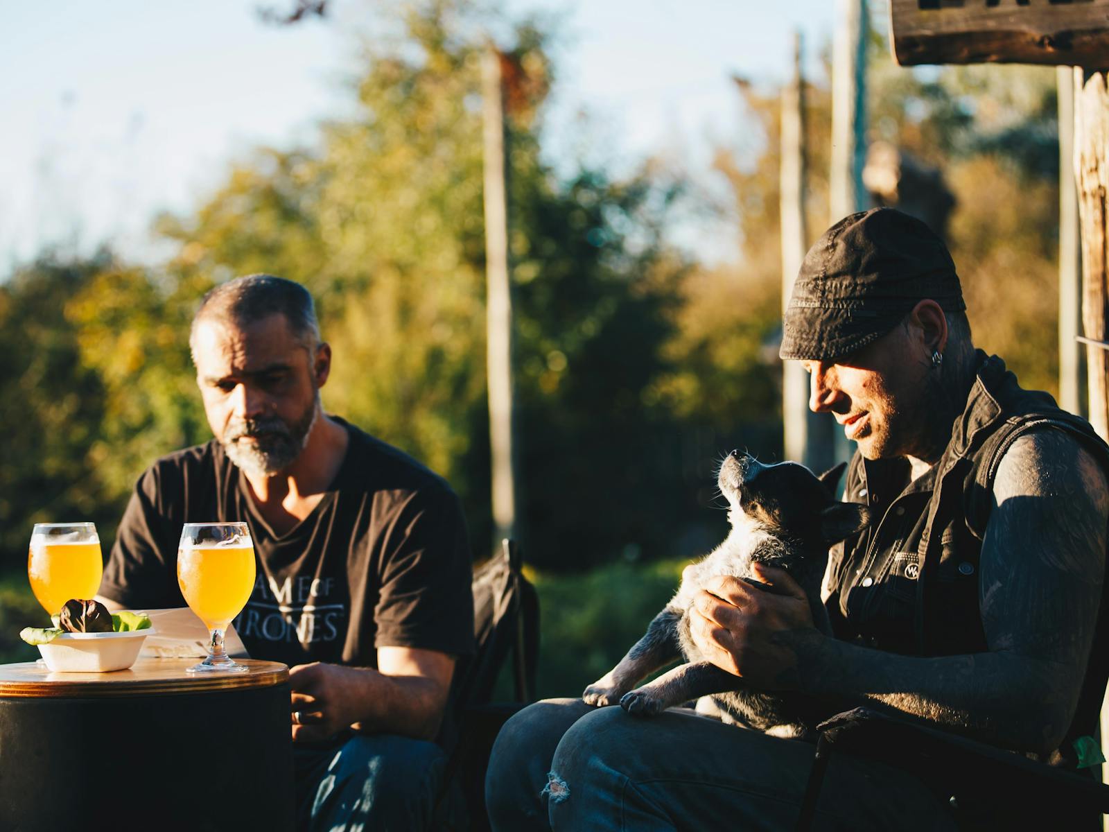Two men sitting outside with glasses of beer and a blue heeler puppy.
