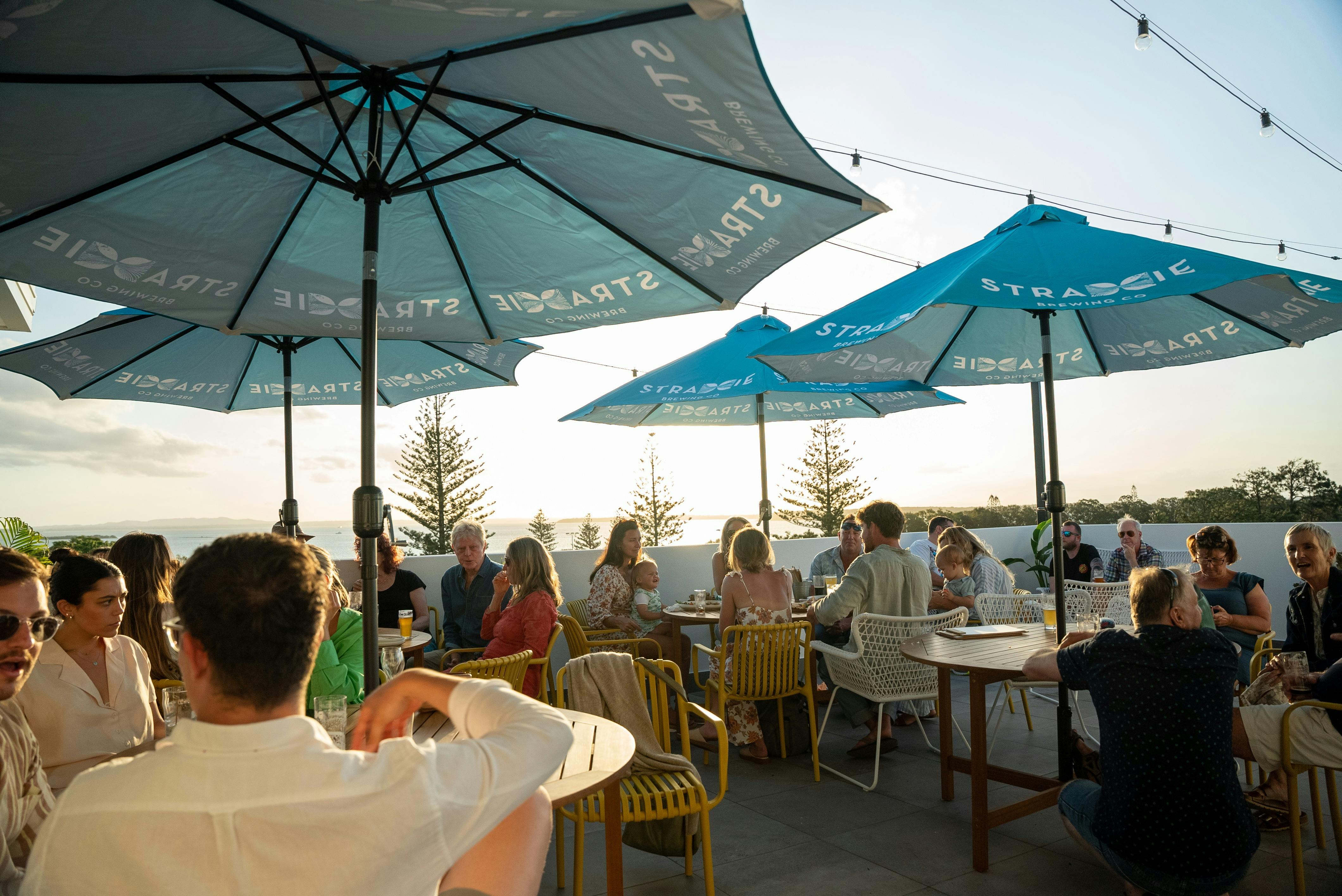 Collection of people seated on yellow chairs, around 4 tables cover by Blue umbrellas