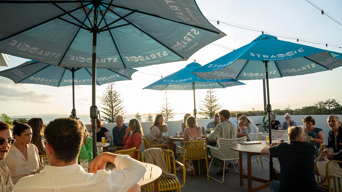 Collection of people seated on yellow chairs, around 4 tables cover by Blue umbrellas