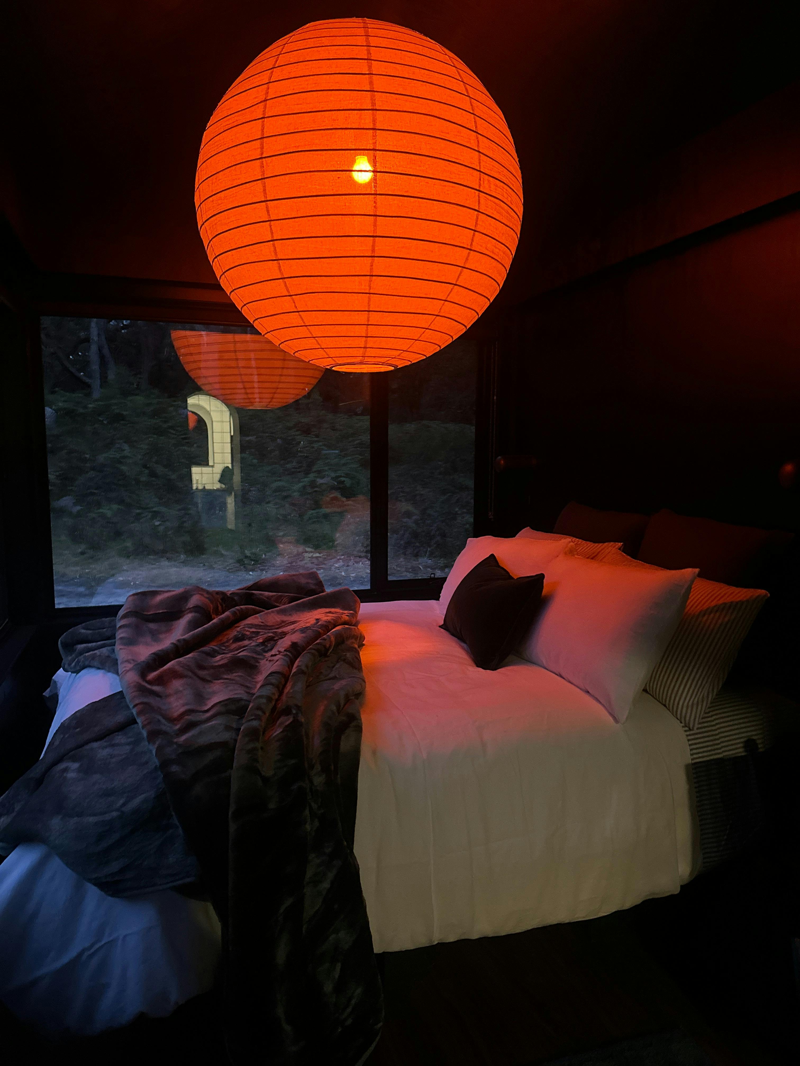 A glowing orange pendant light hangs low over a luxury bed that looks out over native bush
