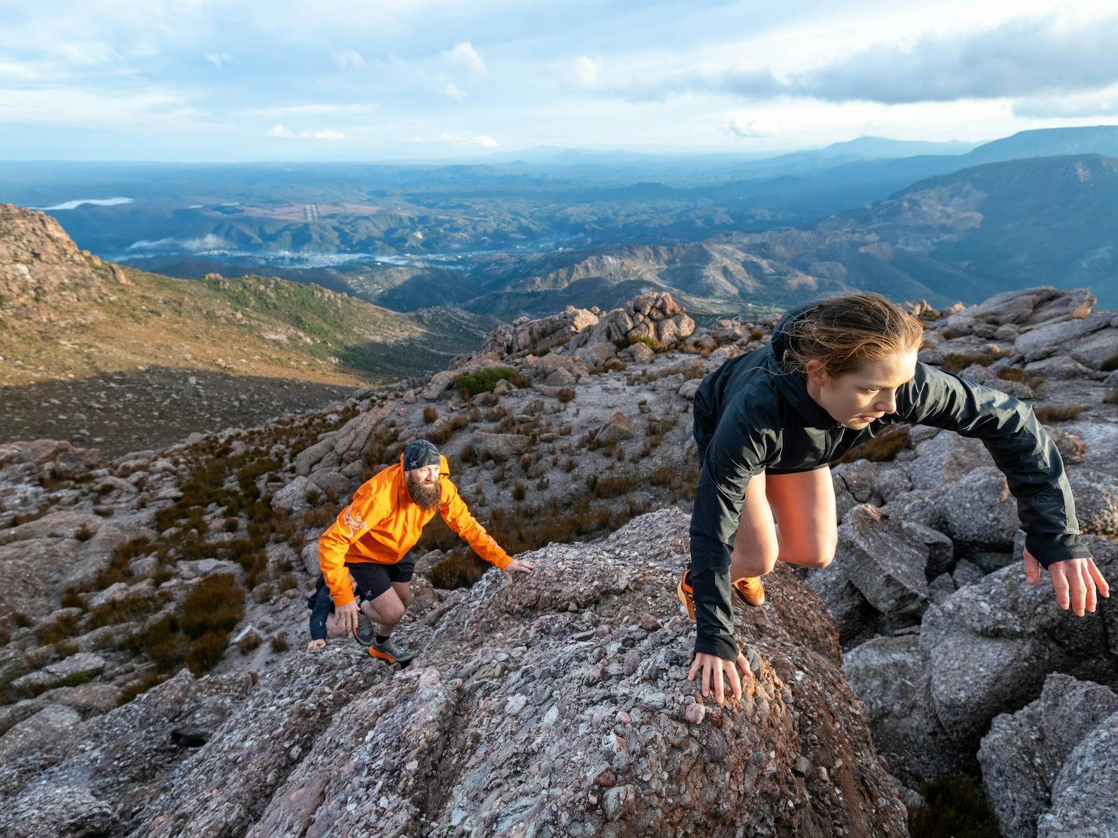 A female and male runner use their hands to climb up the mountain with Queenstown in the distance