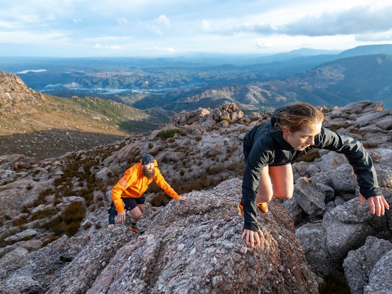 A female and male runner use their hands to climb up the mountain with Queenstown in the distance