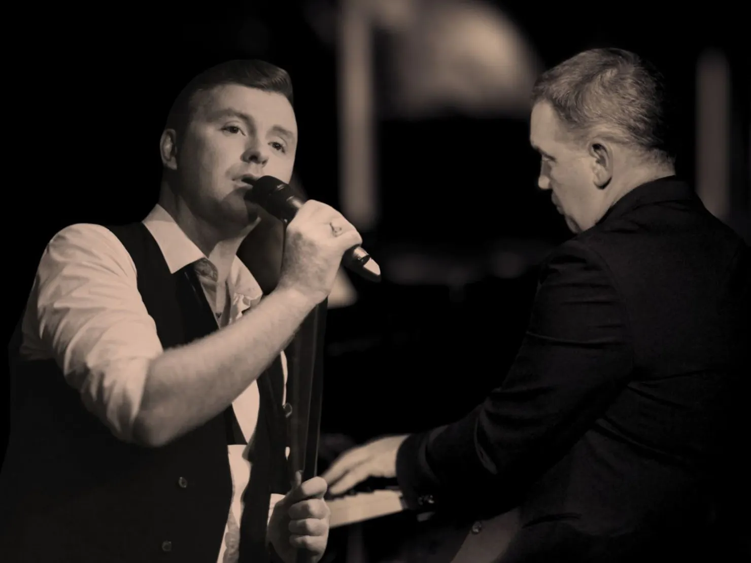 Black and white photo man singing with microphone to mouth and man playing piano in background