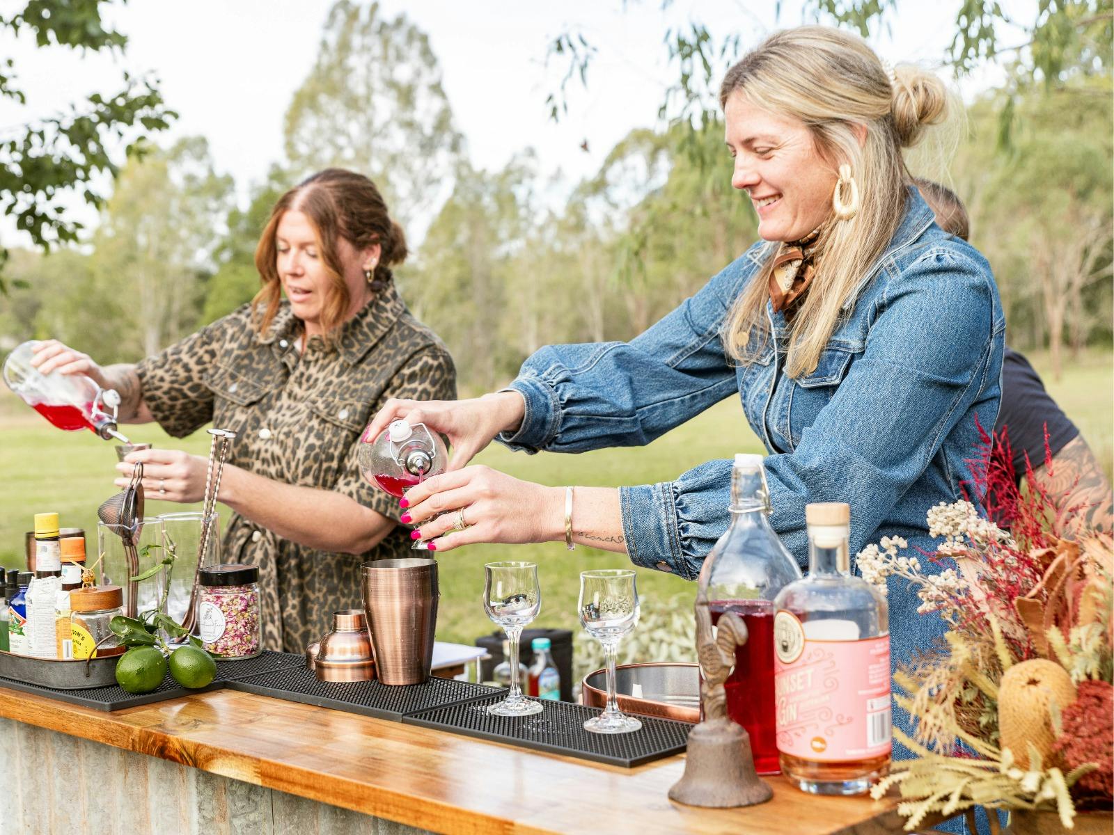 Two women holding syrup bottles pouring them into a cocktail shaker to make cocktails.