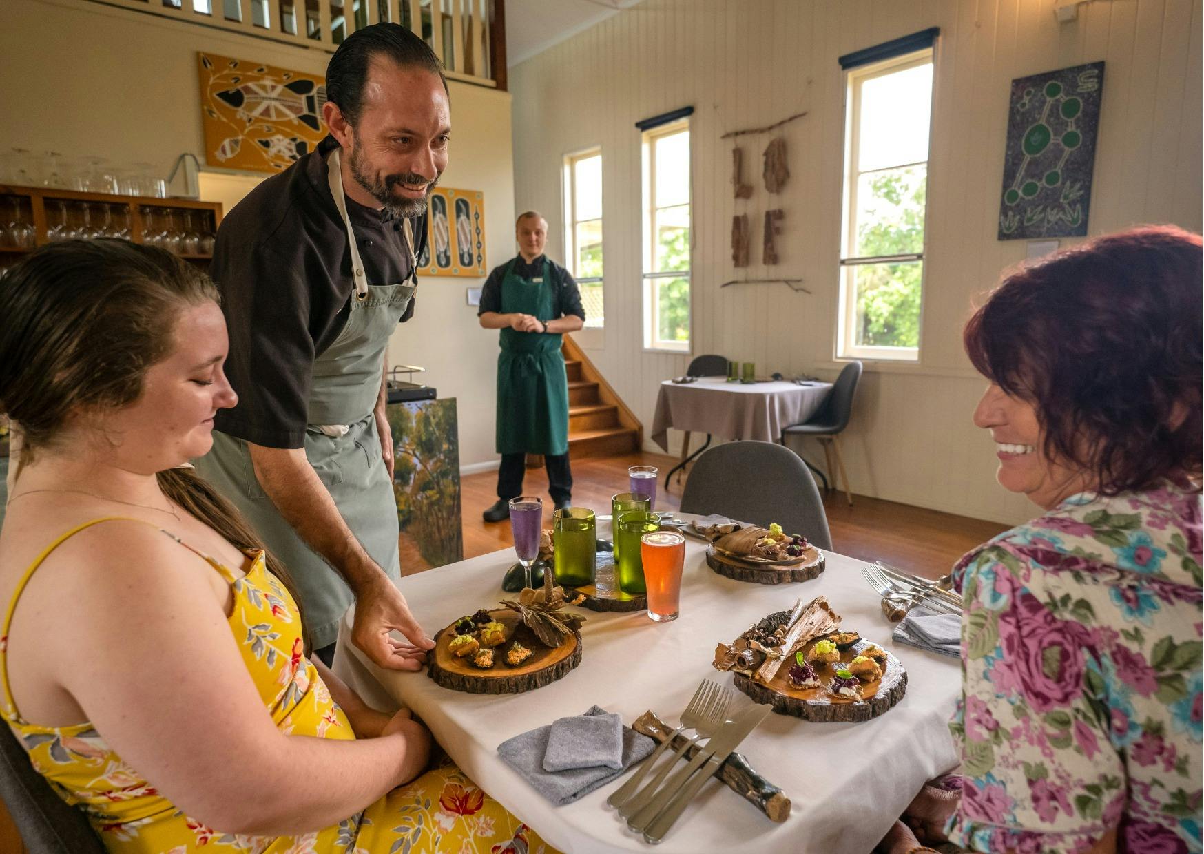 Chef Nik serves two smiling female guests. Three canapes are presented on a polished wood round.