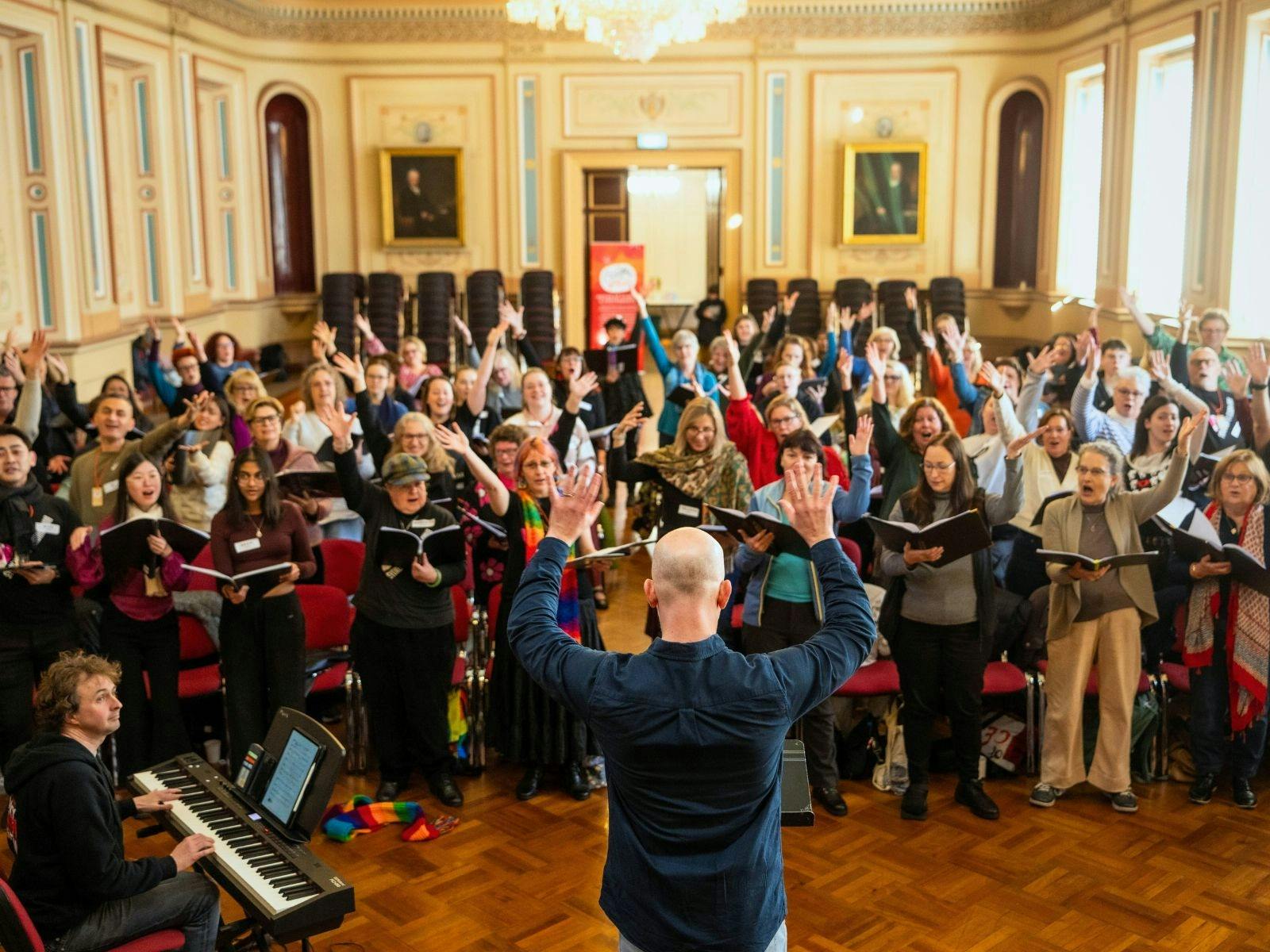Large choir singing in a hall with hands raised, led by a conductor facing away