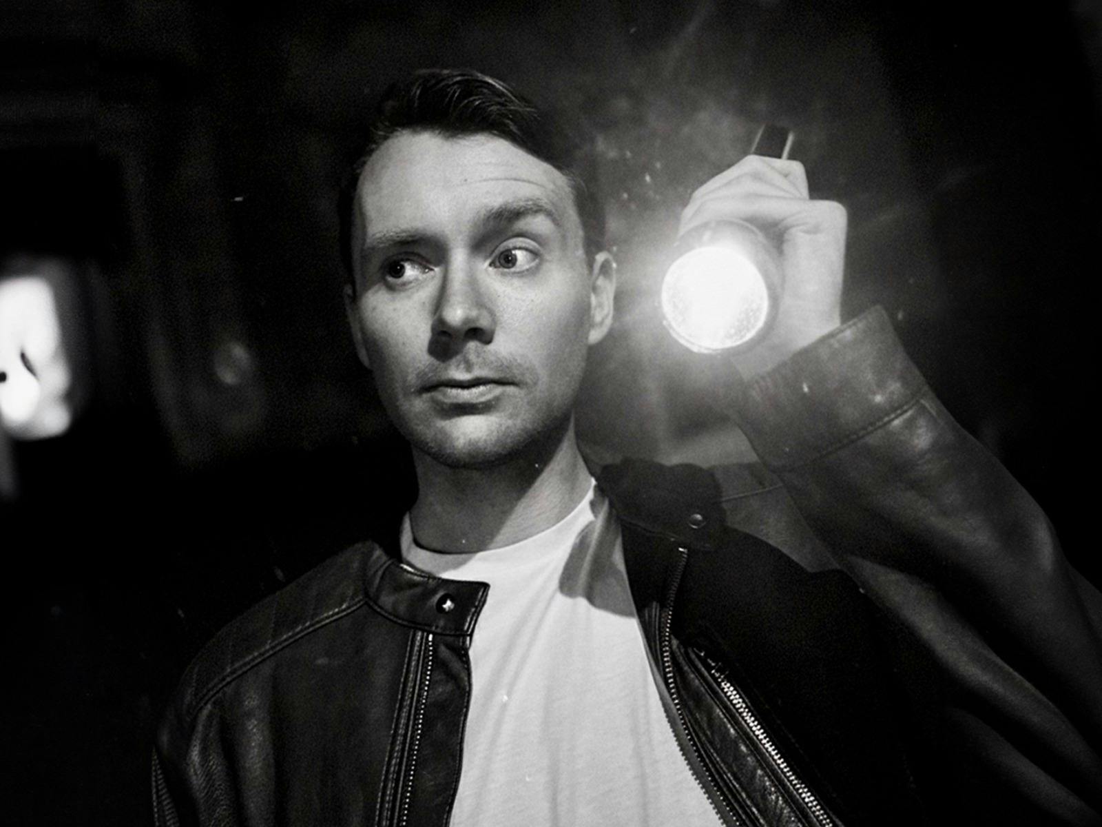 Man wearing a black leather jacket and a white t-shirt holding a torch looking for clues