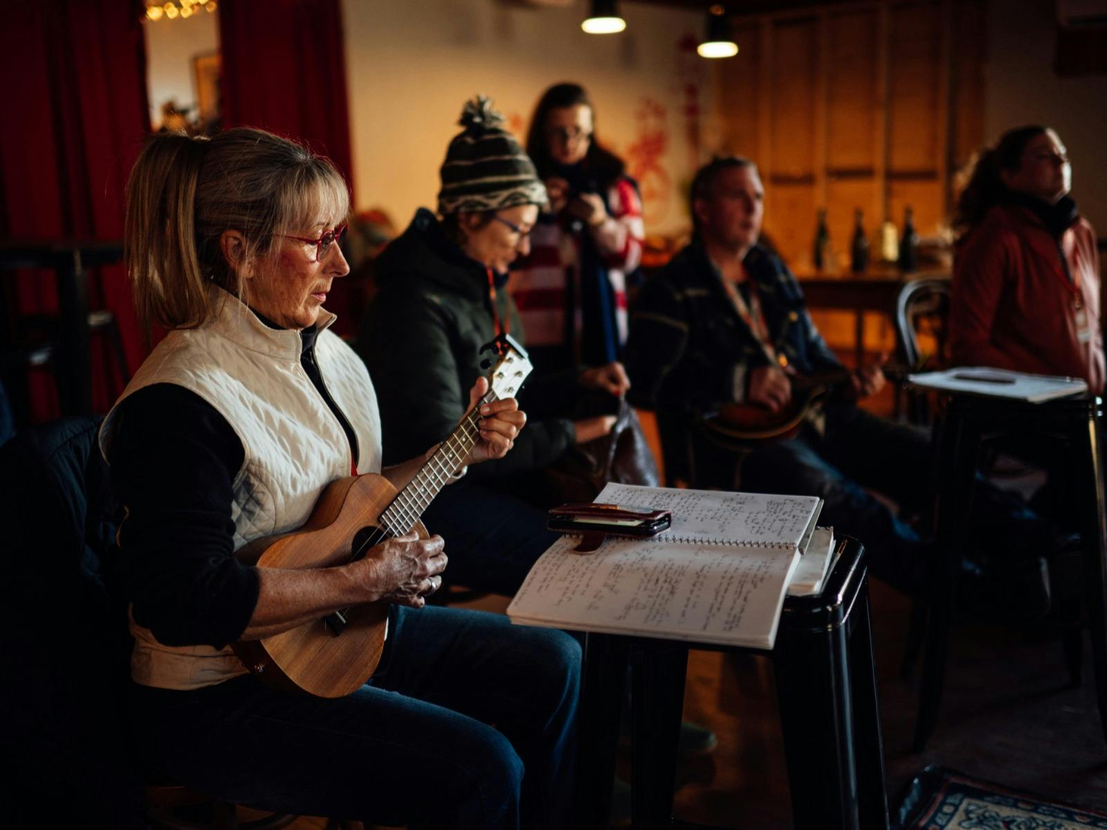 A woman playing a ukulele with a notebook of lyrics open on a stand next to her.