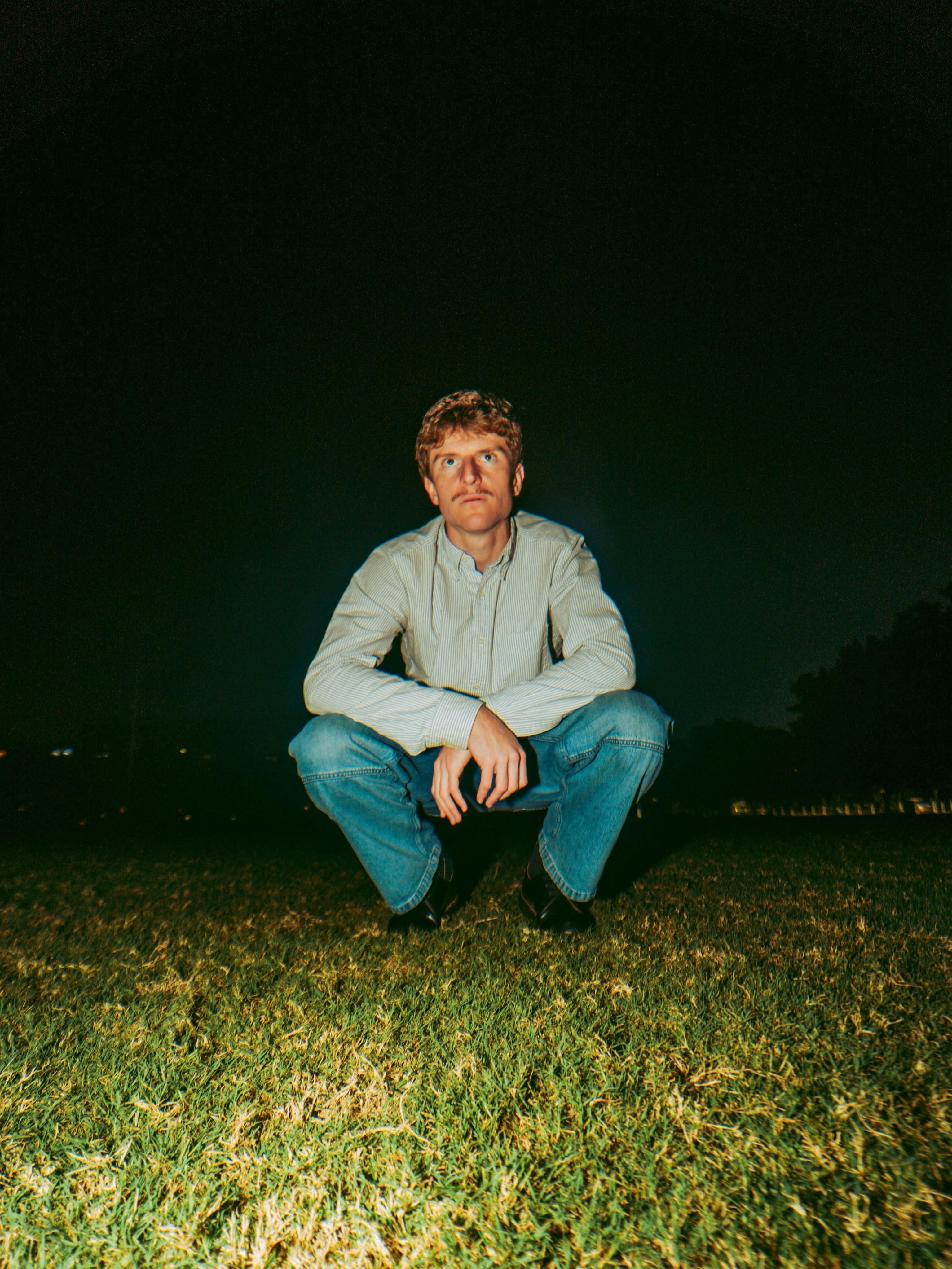 picture of sam squatting on grass wearing jeans and button up shirt