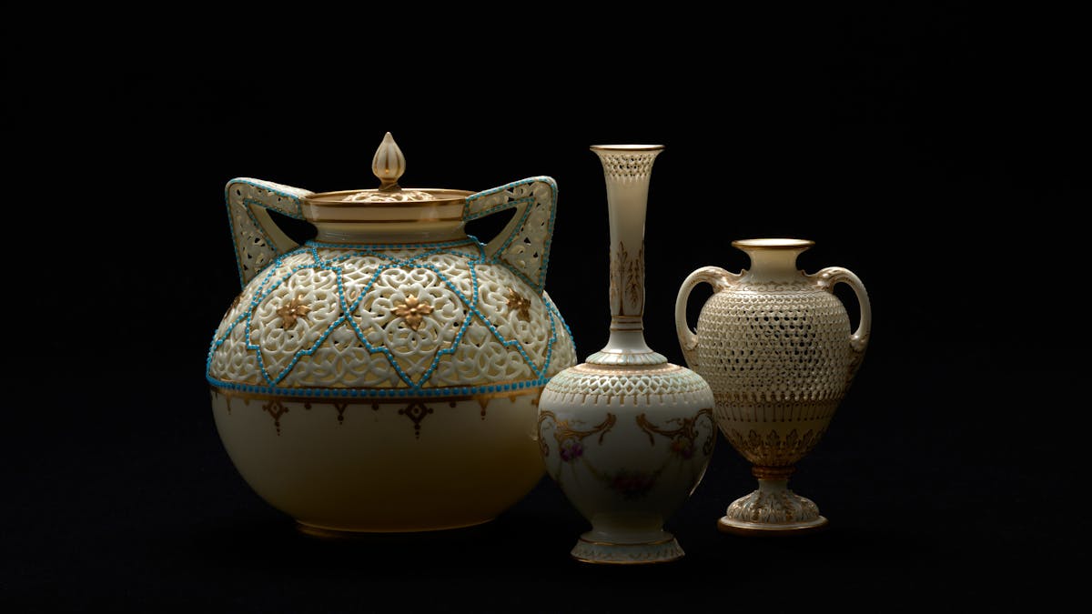A spotlight illuminates three delicate vases and urns with gold gilt detail A spotlight illuminates three delicate vases and urns with gold gilt detail
