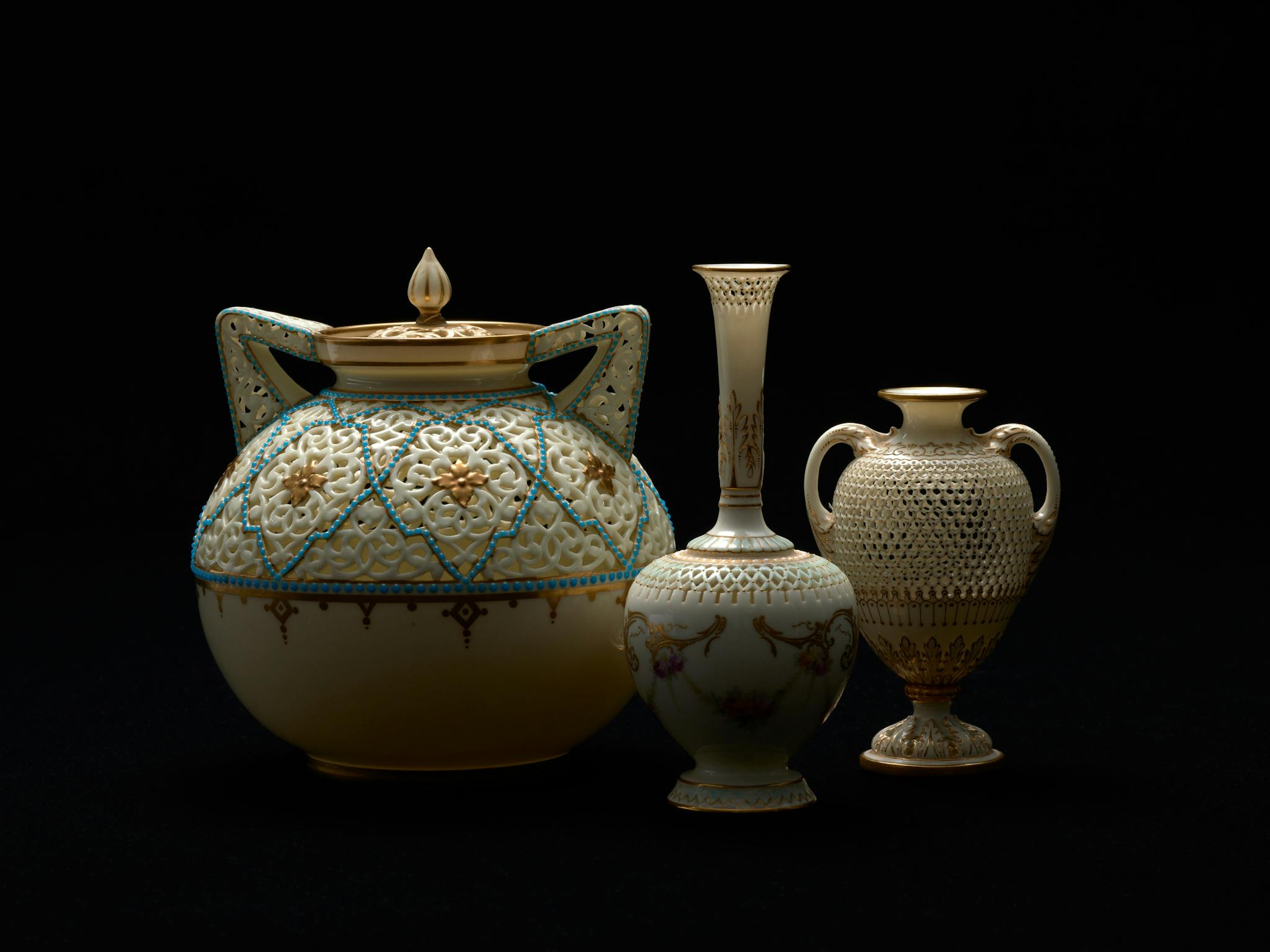 A spotlight illuminates three delicate vases and urns with gold gilt detail A spotlight illuminates three delicate vases and urns with gold gilt detail