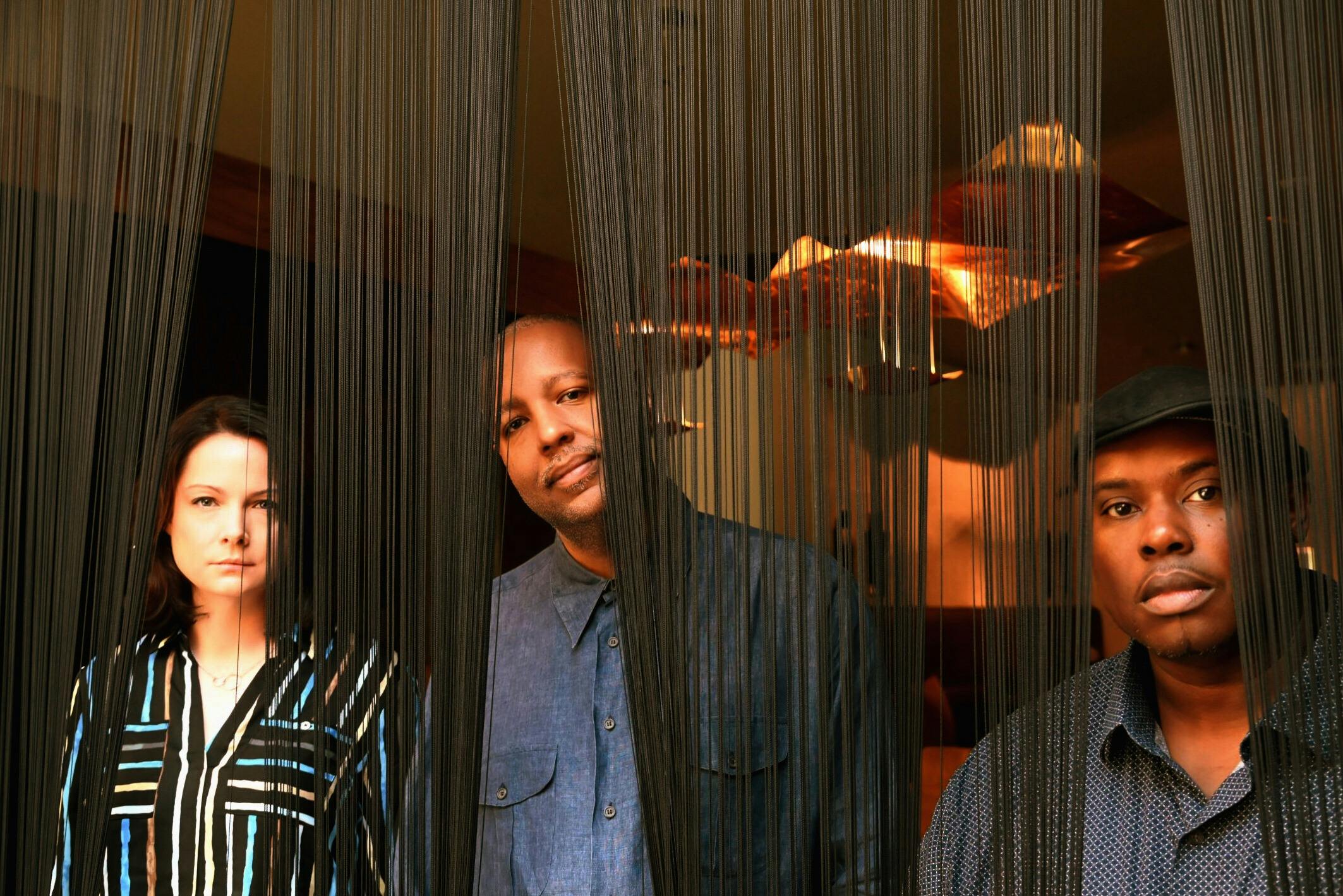 Kris Davis Trio, one woman and two men pose behind fringe curtain