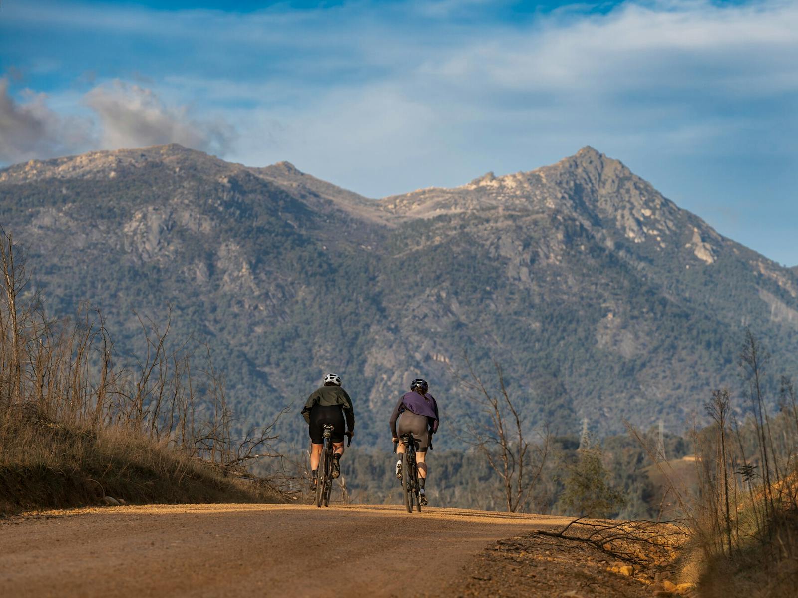 The back of two people on bikes riding on a gravel road with large mountain in backdrop.