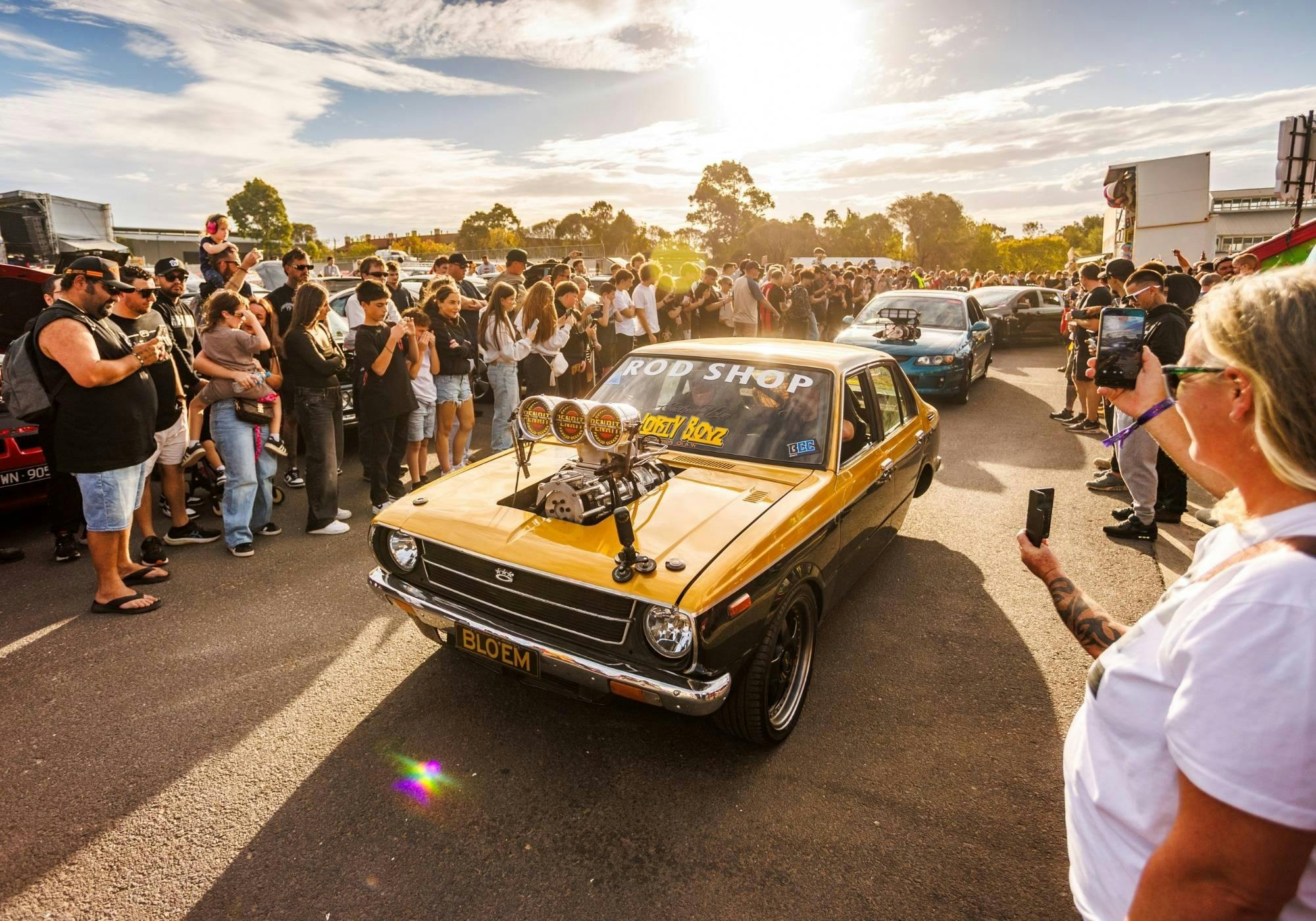 Gold classic sedan with an exposed supercharger cruising between crowds in the afternoon sun.