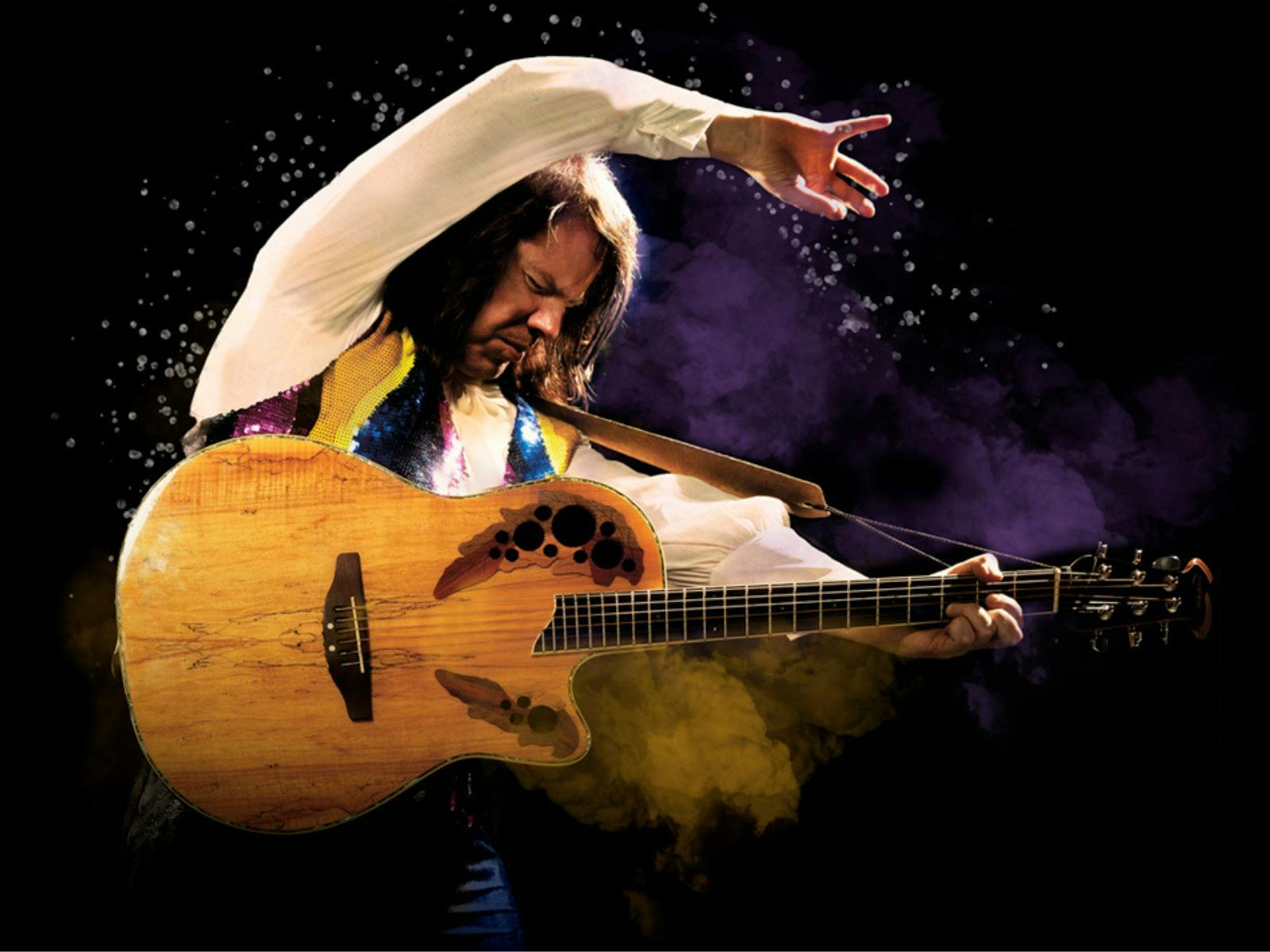 A musician with long hair passionately strums an acoustic guitar, with one arm raised