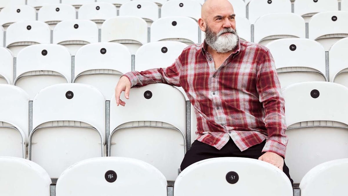 A bearded adult wearing a red plaid shirt sits alone among rows of light grey stadium-style seats