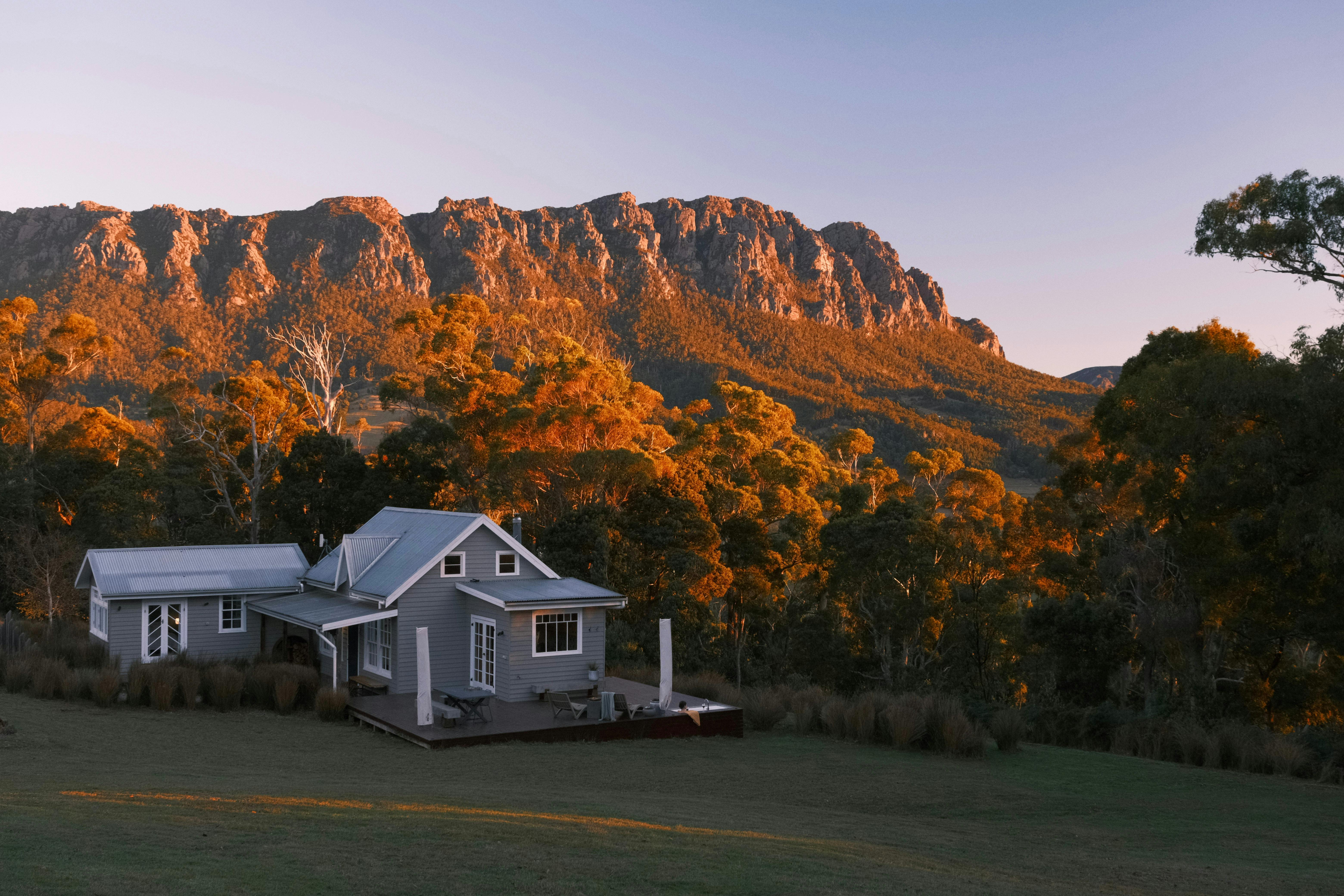 A grey cabin with forest in the background and the setting sun on a large mountain