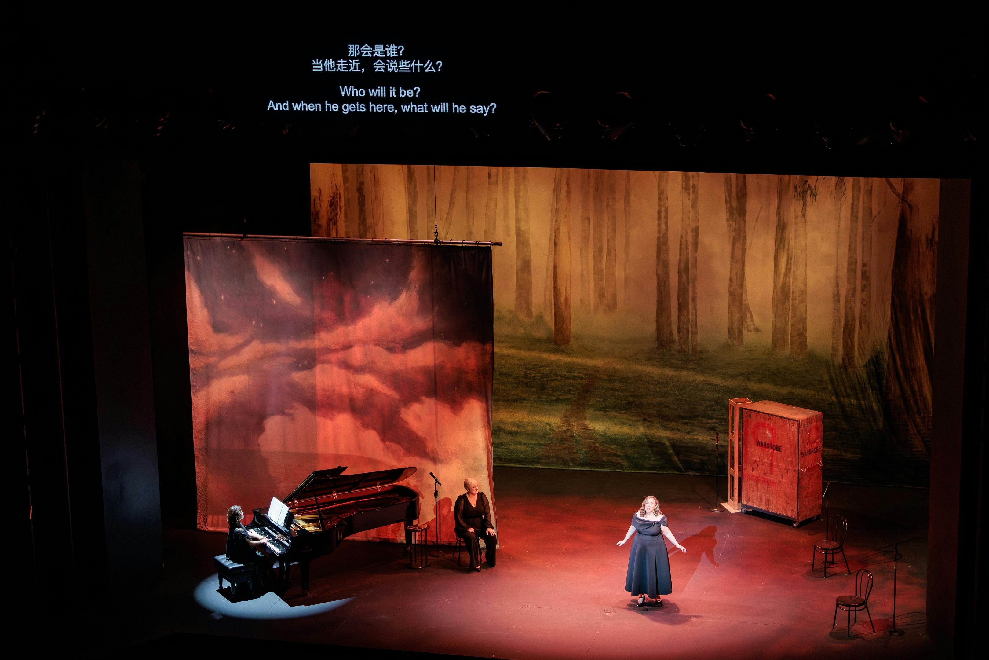An opera singer and pianist perform on a stage with a surtitle screen seen above it