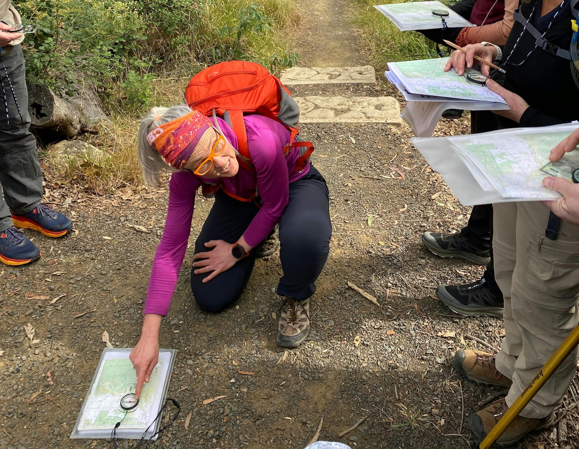 Woman in pink shirt and red backpack is kneeling on the ground and pointing to a compass on a map