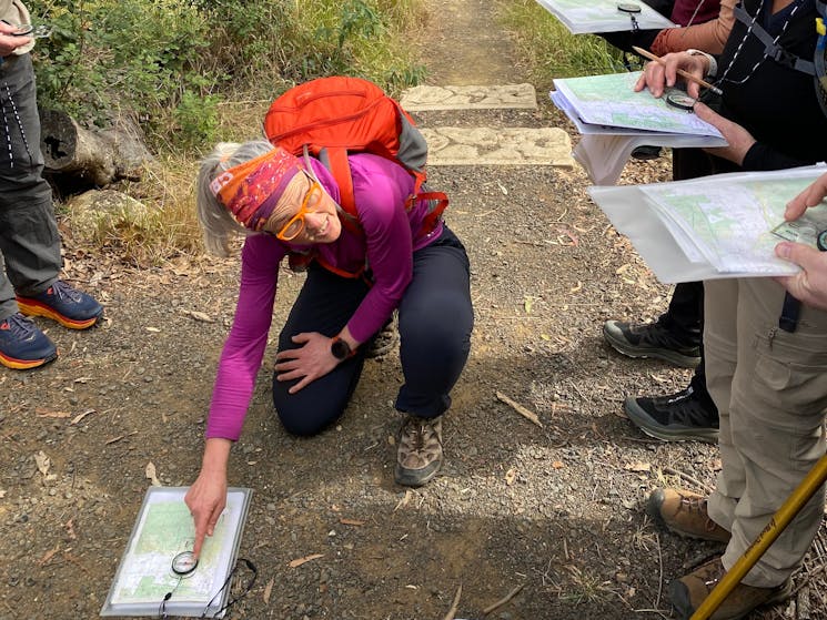 Woman in pink shirt and red backpack is kneeling on the ground and pointing to a compass on a map