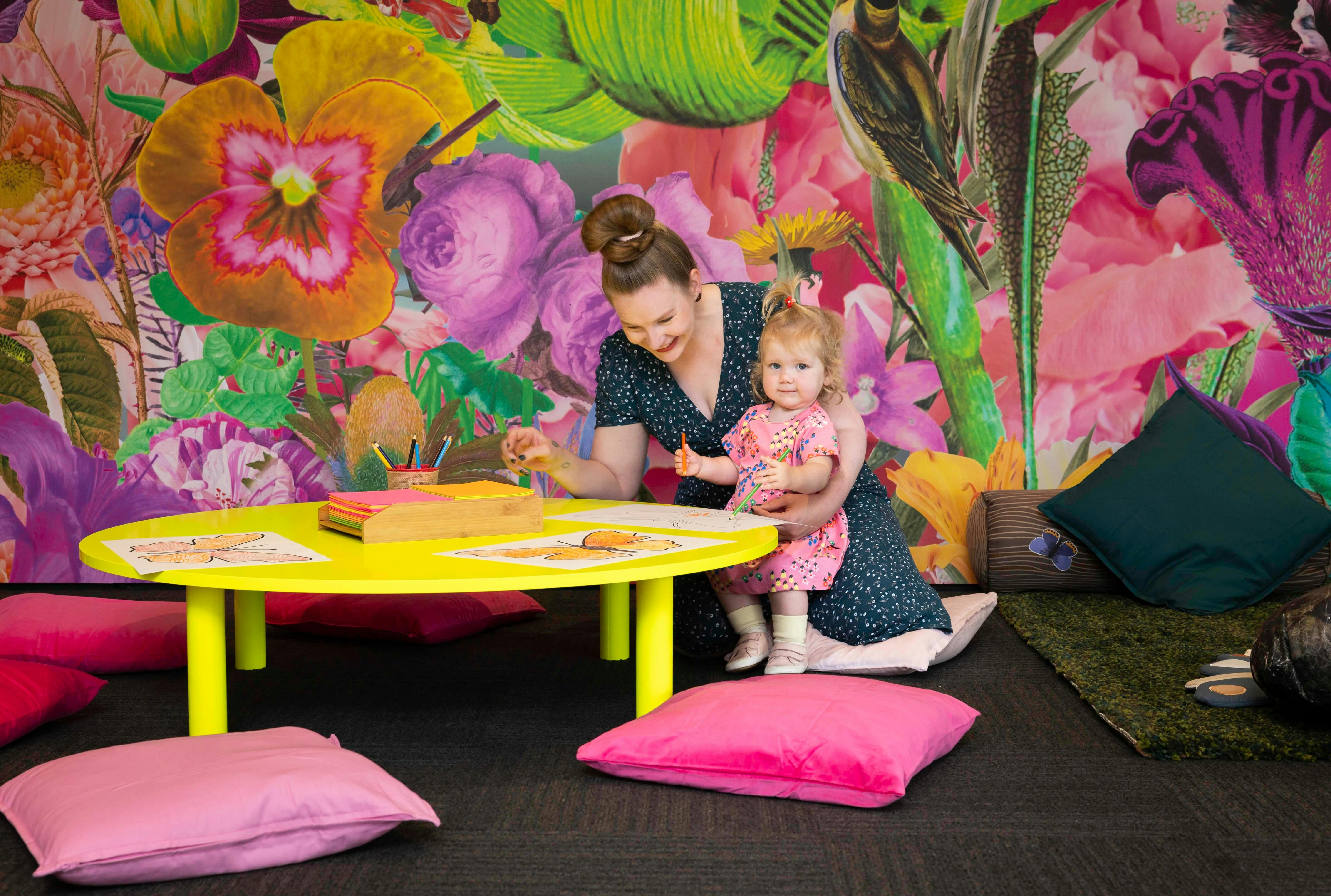 A picture of a woman and a child drawing on a fluorescent yellow table in front of a colourful wall