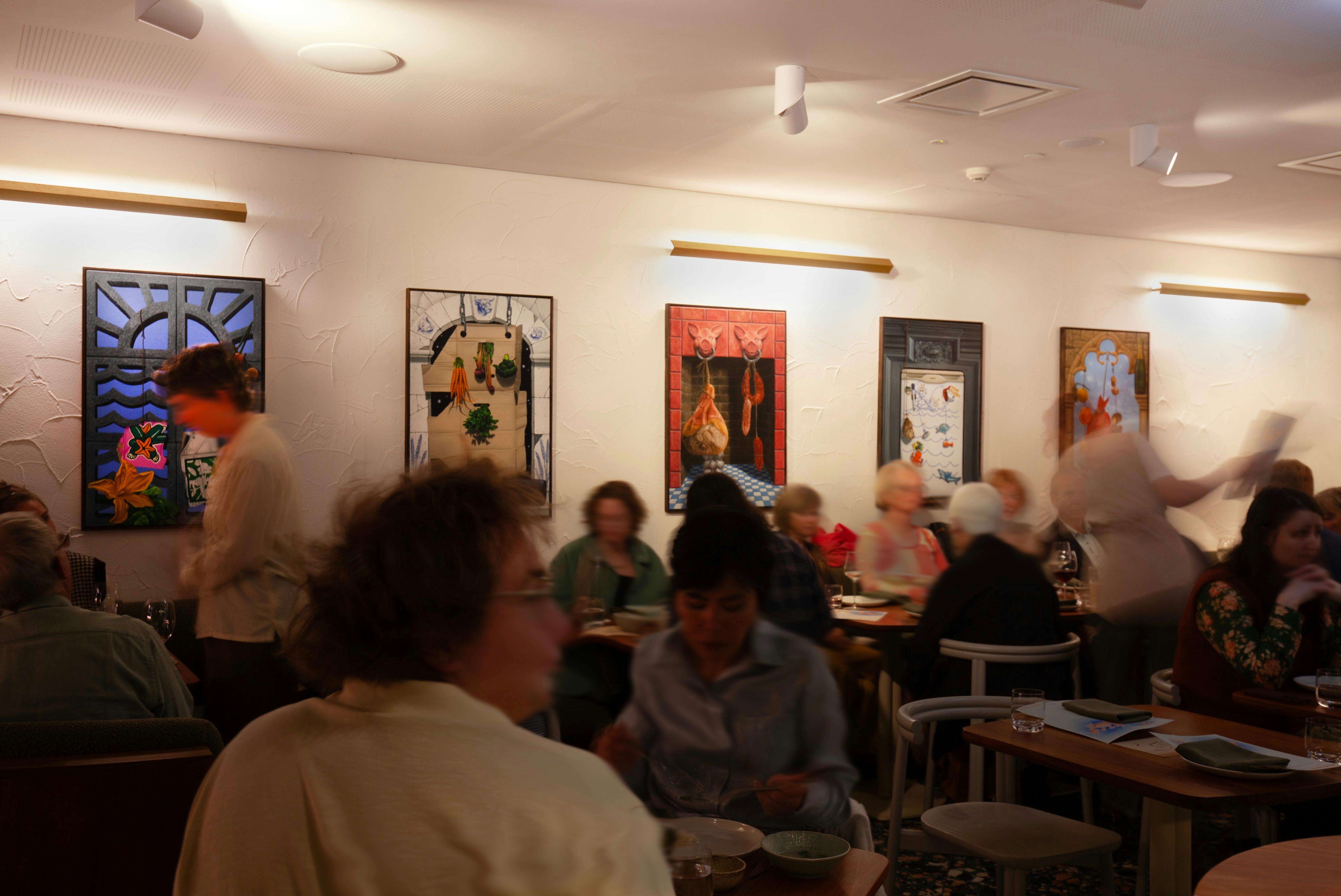 A restaurant full of people moving, with colourful paintings hung on the walls.