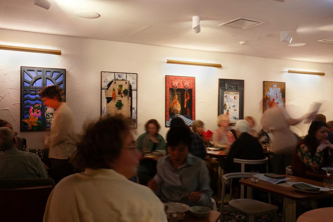 A restaurant full of people moving, with colourful paintings hung on the walls.