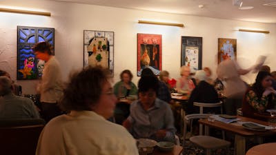 A restaurant full of people moving, with colourful paintings hung on the walls.