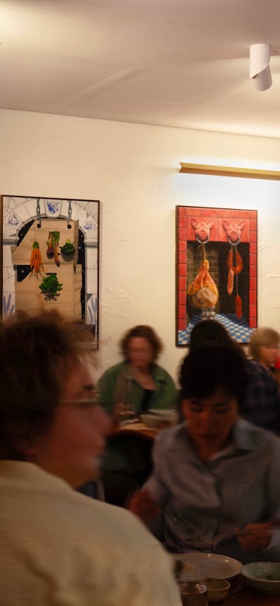 A restaurant full of people moving, with colourful paintings hung on the walls.
