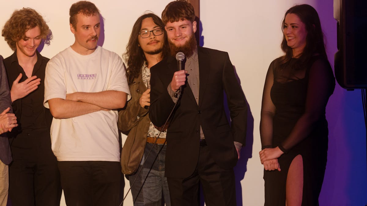 A group of five smiling individuals stand on a stage. One man in a suit holds a microphone.