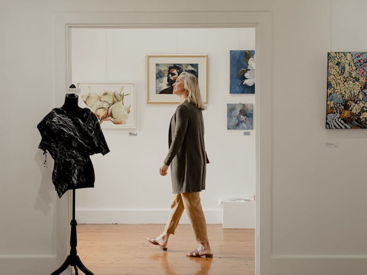A woman is seen through the doorway of a white art gallery, paintings on the walls around her.
