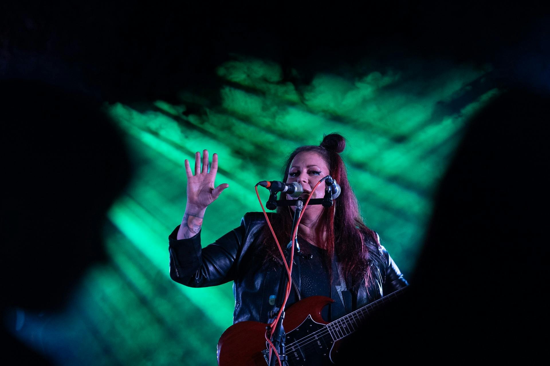 A female musician performs on stage under dramatic green lighting. She wears a black leather jacket