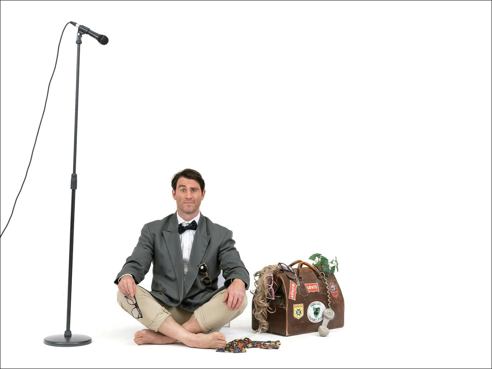 A man in a tuxedo shirt and jacket with shorts sitting on the ground between a mic and a suitcase A man in a tuxedo shirt and jacket with shorts sitting on the ground between a mic and a suitcase