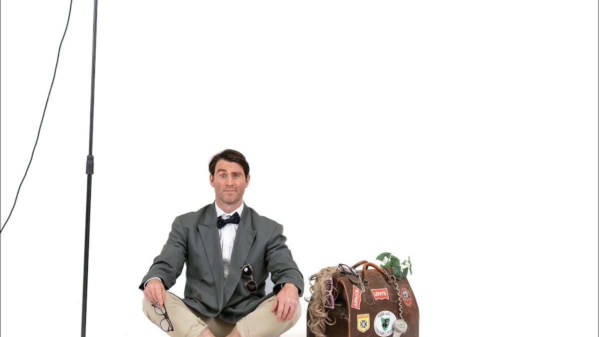 A man in a tuxedo shirt and jacket with shorts sitting on the ground between a mic and a suitcase