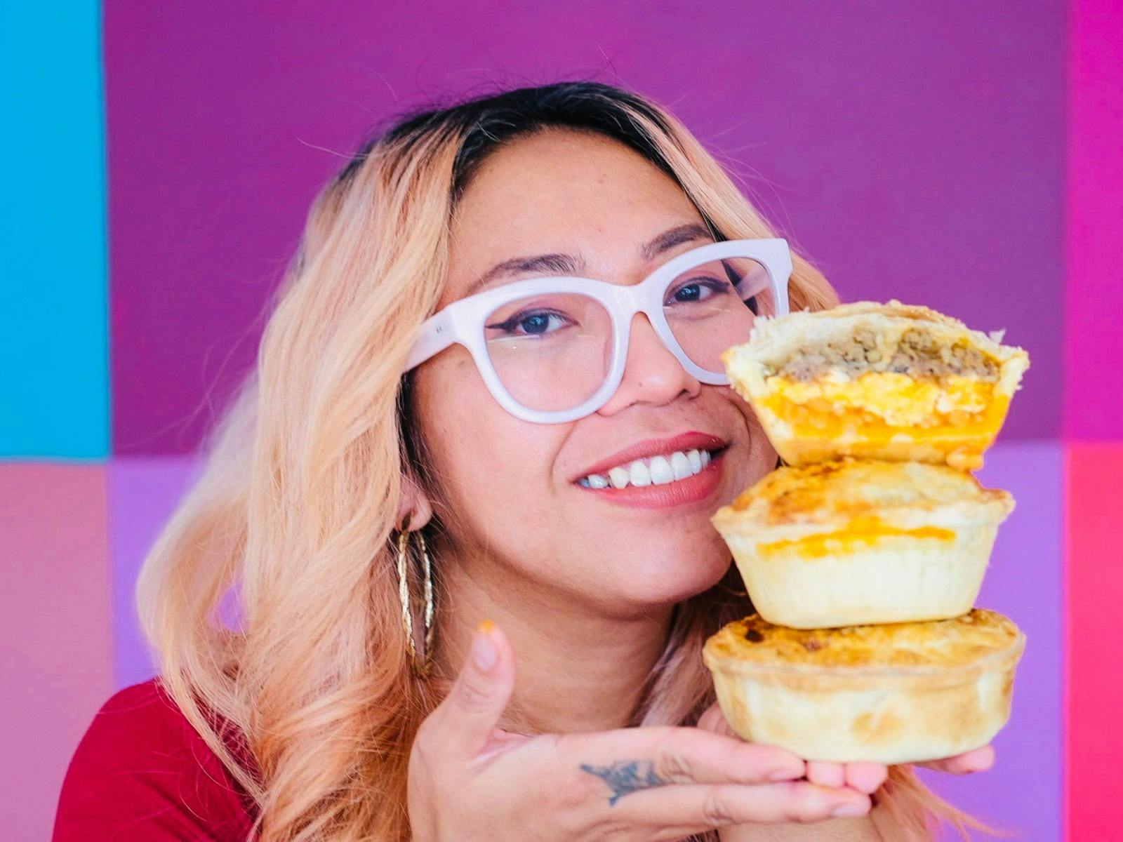Lilly smiling and holding three pies in front of a vibrant pink background