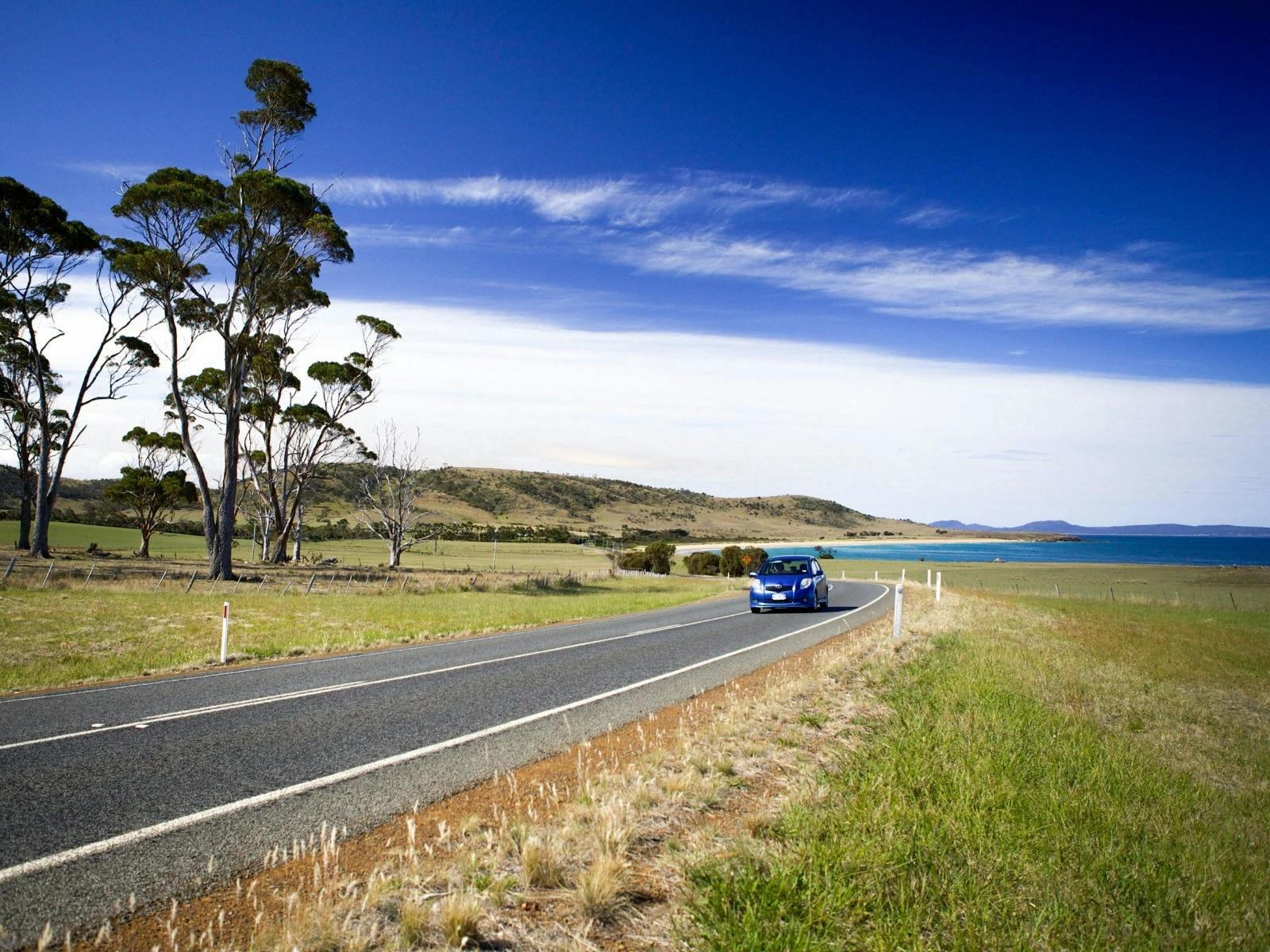 A blue car drives along the e on a clear, sunny day by the beach
