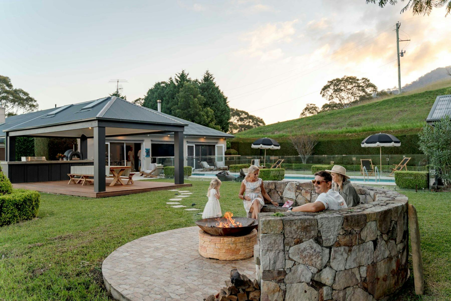 Image of back outdoor area. A family sits around the open firepit. Home sits beautifully in backdrop