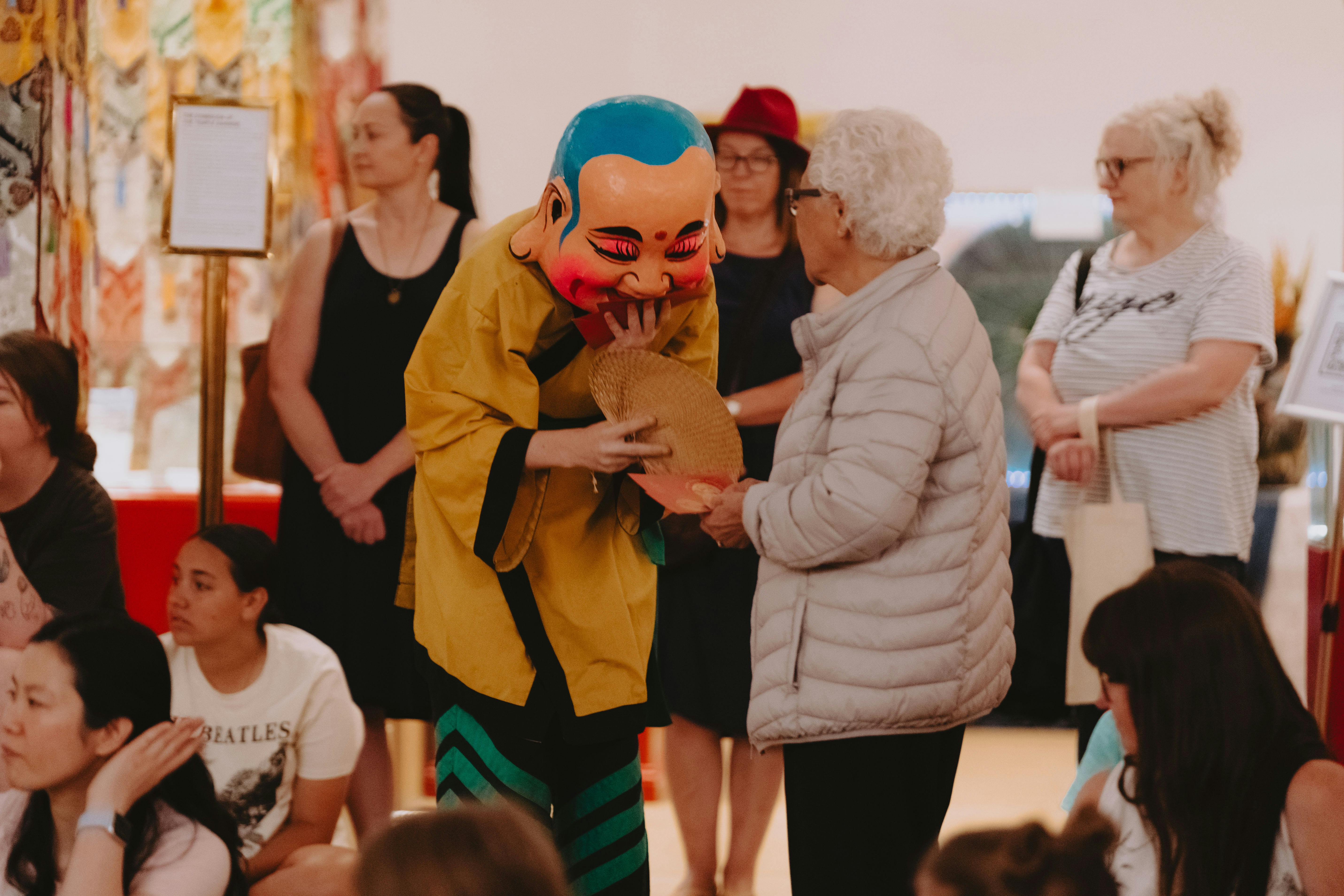 Image of someone in chinese attire and mask accepting offering from a woman in grey jacket