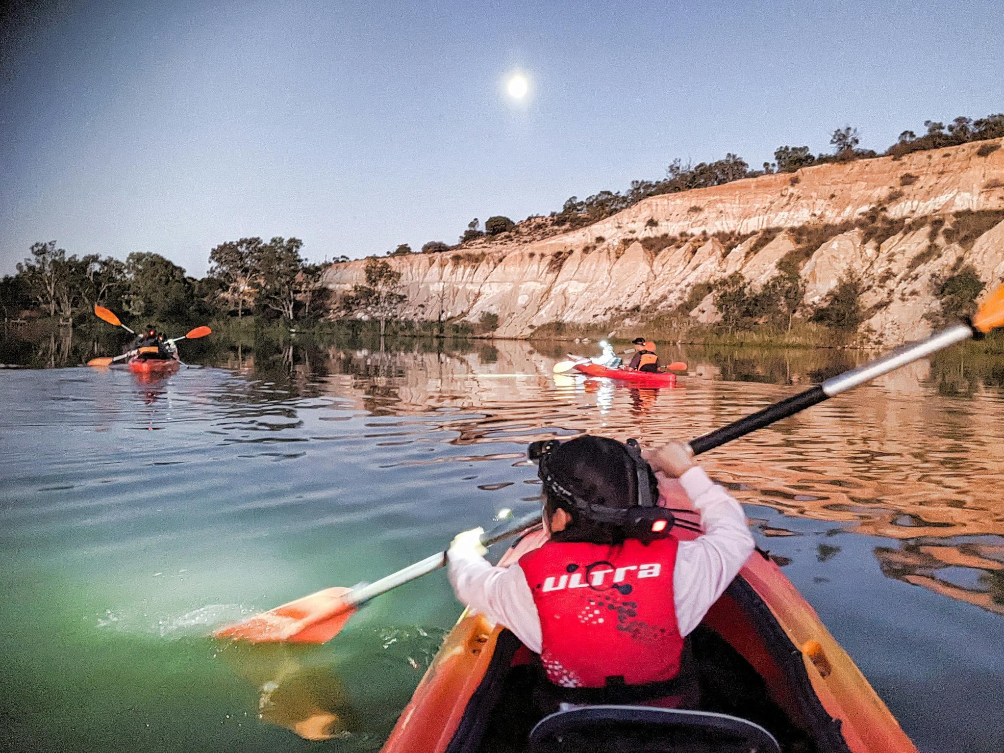 Girl paddling in the front of a double kayak, at dusk, under a full moon, wearing a  head torch.