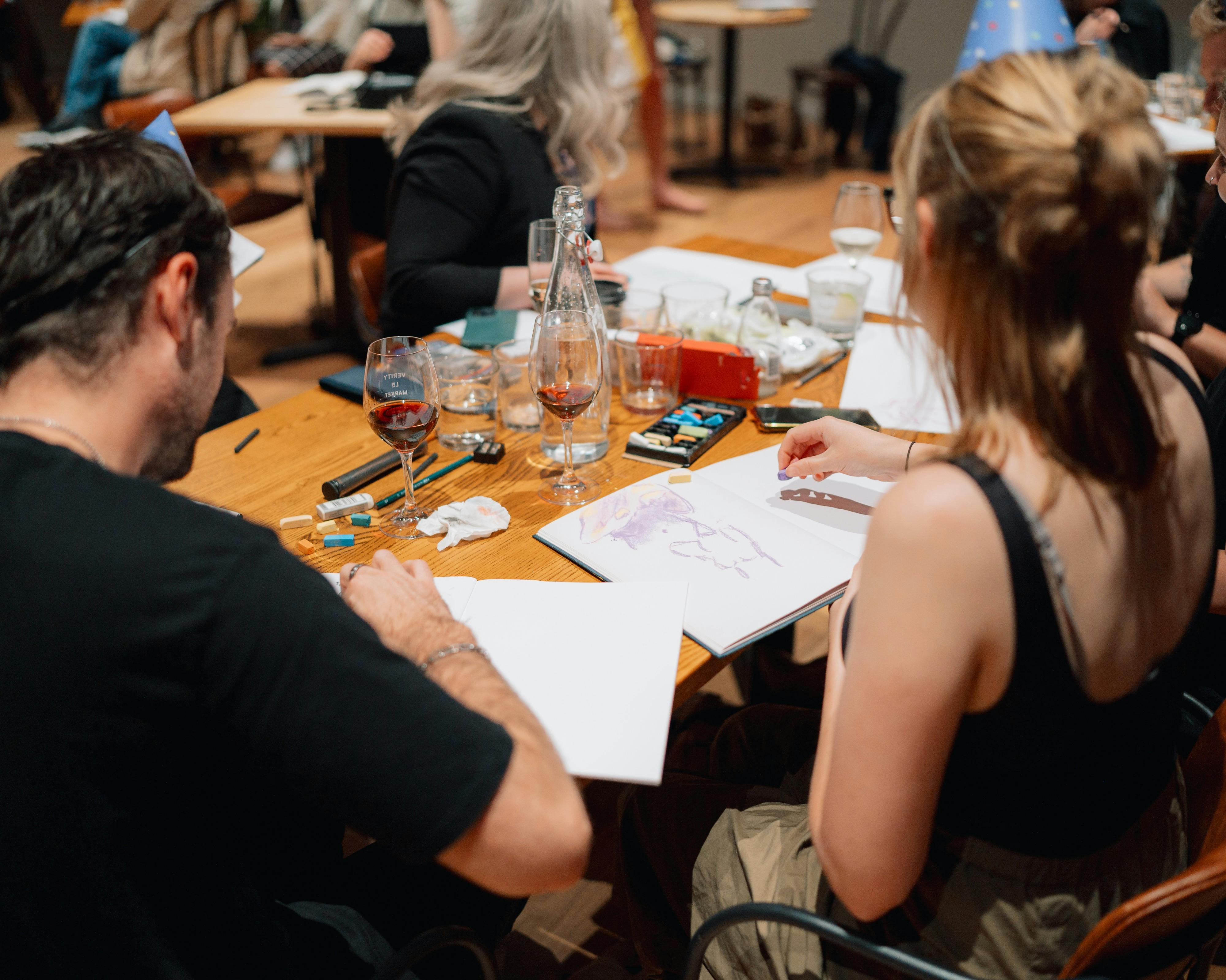 People sitting at a table drawing, with glasses of red wine