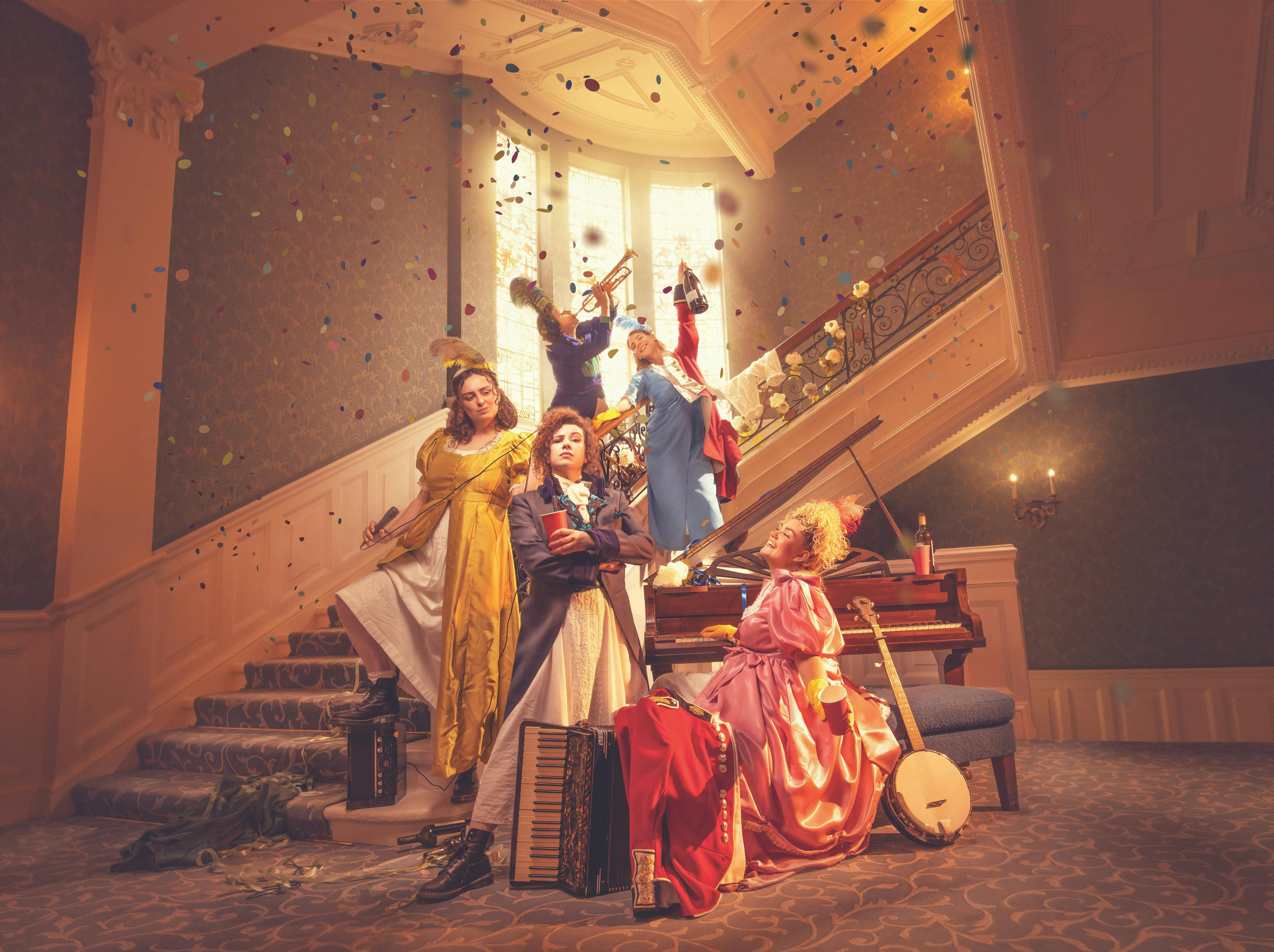 Four women in colorful period costumes pose on a grand staircase, surrounded by musical instruments
