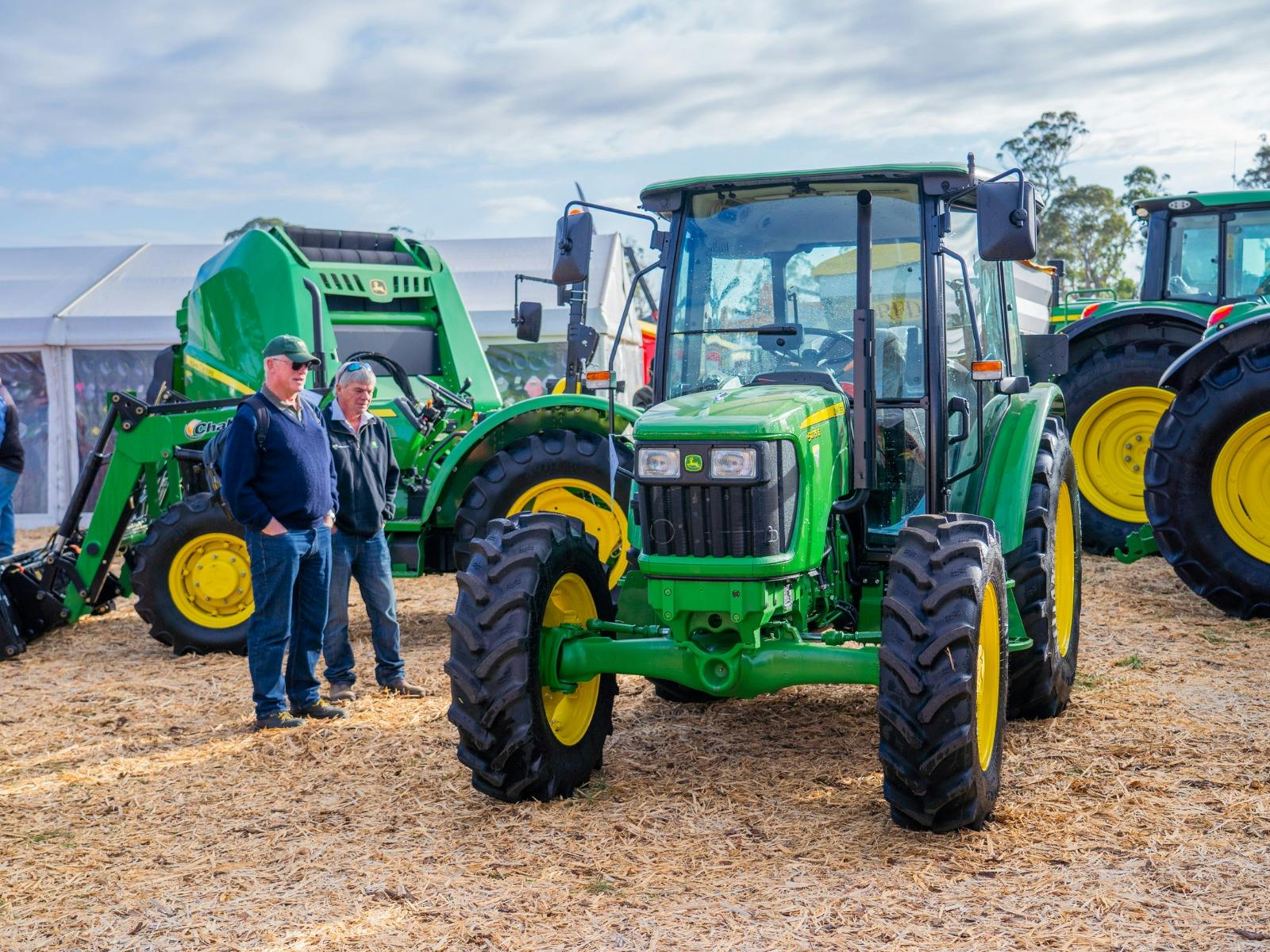 Two men stand beside a John Deere tractor, smiling with exhibitor stalls in the background.